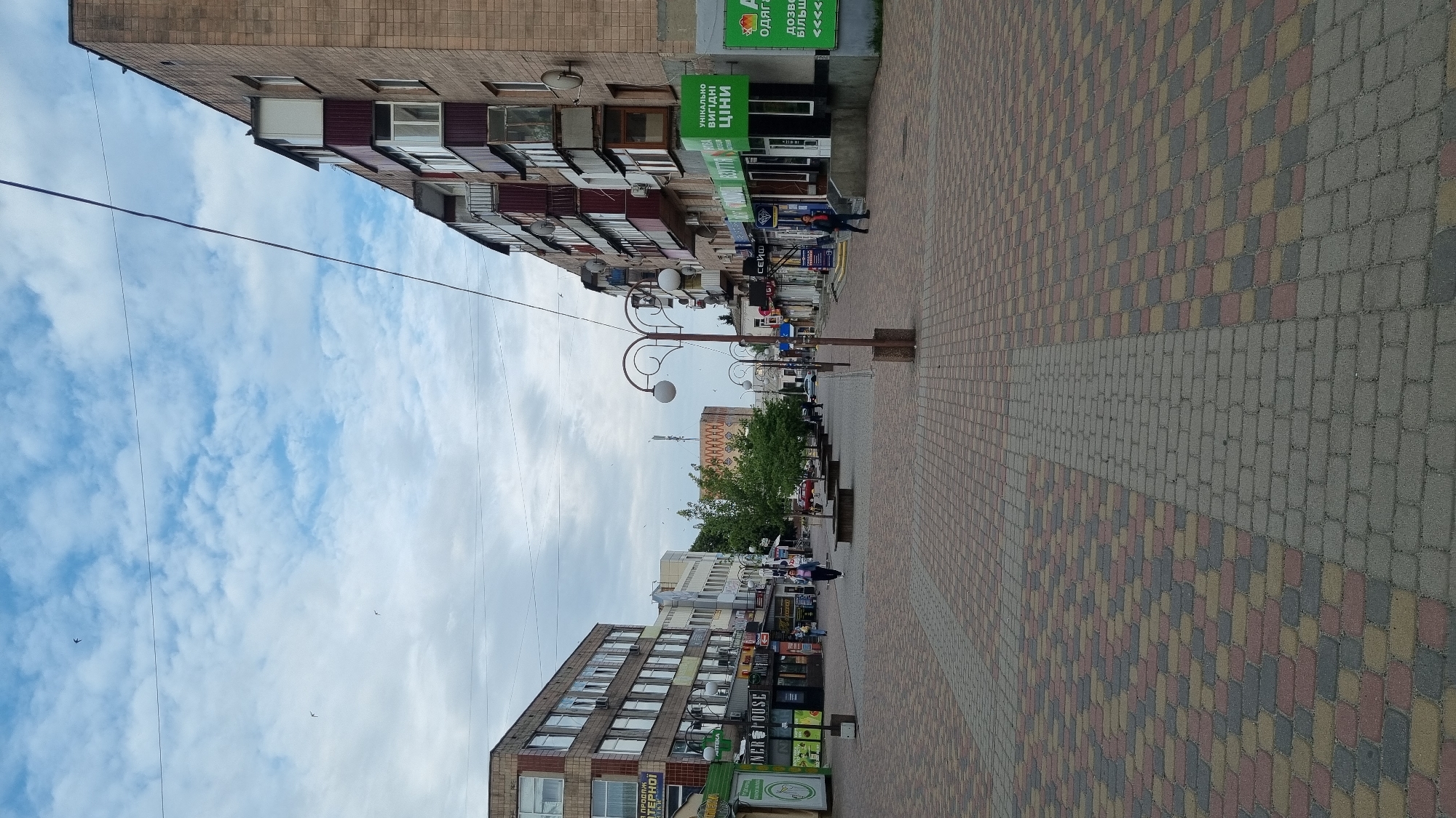 The image depicts a street scene in the city of Smila, Ukraine. The foreground is dominated by a pedestrian walkway paved with multi-colored bricks in a repeating pattern of red, yellow, and gray. The walkway stretches into the distance, flanked by buildings on both sides. The left side of the street features a multi-story building with numerous windows and signs for businesses. On the right side, another multi-story residential building with balconies is visible, with ground-floor commercial establishments below. Several street lamps with spherical lights line the walkway. Trees are planted in rectangular beds along the pedestrian area. The sky above is cloudy with patches of blue and several birds are in flight. There are a few individuals walking on the pedestrian path, and vehicles are parked along the street in the background. The overall impression is a moderately busy urban commercial and residential street.