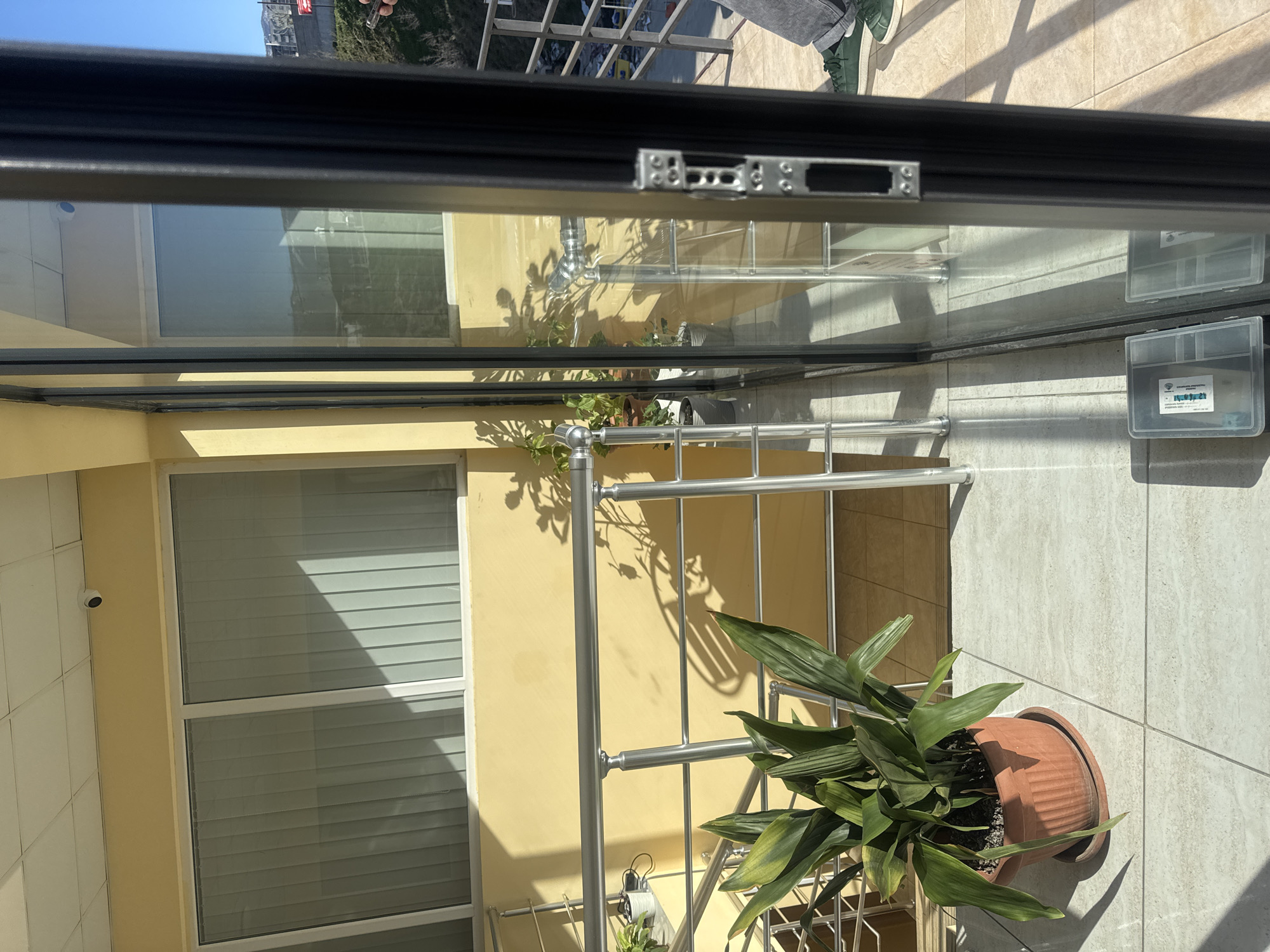 Here's a concise description of the scene:
The image shows a view from inside a building, likely an apartment or office in Tbilisi, Georgia, looking out onto a balcony. The vantage point is through a partially opened window or door. The exterior includes a yellow wall with a window, a security camera, a metal railing, a potted plant with large green leaves. The balcony is tiled. Reflections in the window show a view of the city. There is a plastic container attached on the glass.