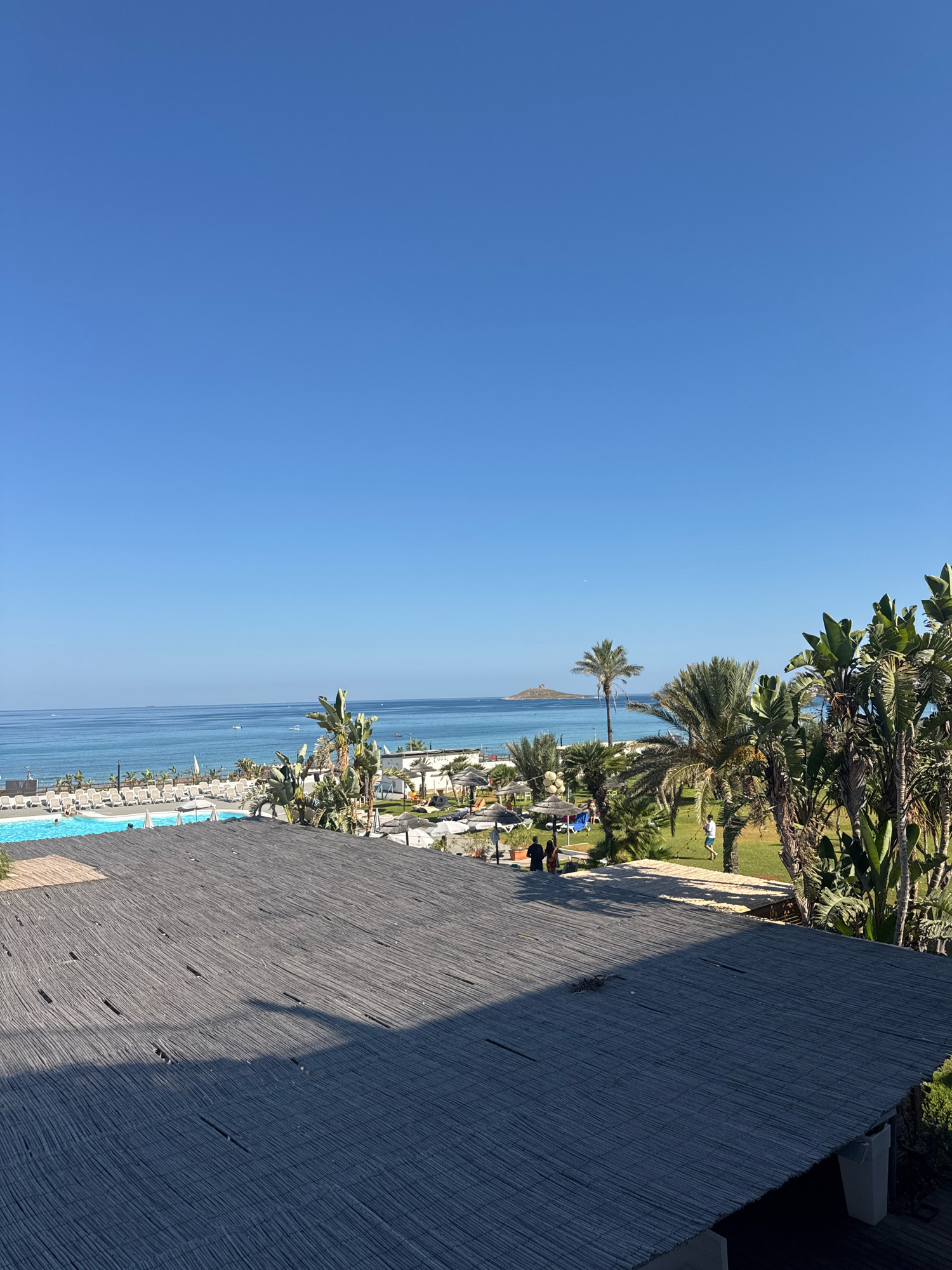 The image shows a view from a rooftop looking out towards the sea. In the foreground, a dark, thatched-like roof covers the majority of the frame. Beyond this, a swimming pool with lounge chairs surrounds it, and further out, a beach with more lounge chairs and umbrellas is visible. Palm trees and lush greenery dot the landscape, with a few people scattered throughout the resort area. The sea stretches out to the horizon, meeting a clear blue sky. In the distance, a small tower-like structure stands on a rocky outcrop in the water. The scene is bathed in bright sunlight, suggesting a warm, clear day. The location appears to be a coastal resort in the city of Isola delle Femmine, Italy, given the characteristic architecture and the presence of the distinctive tower, which is a landmark of the area.