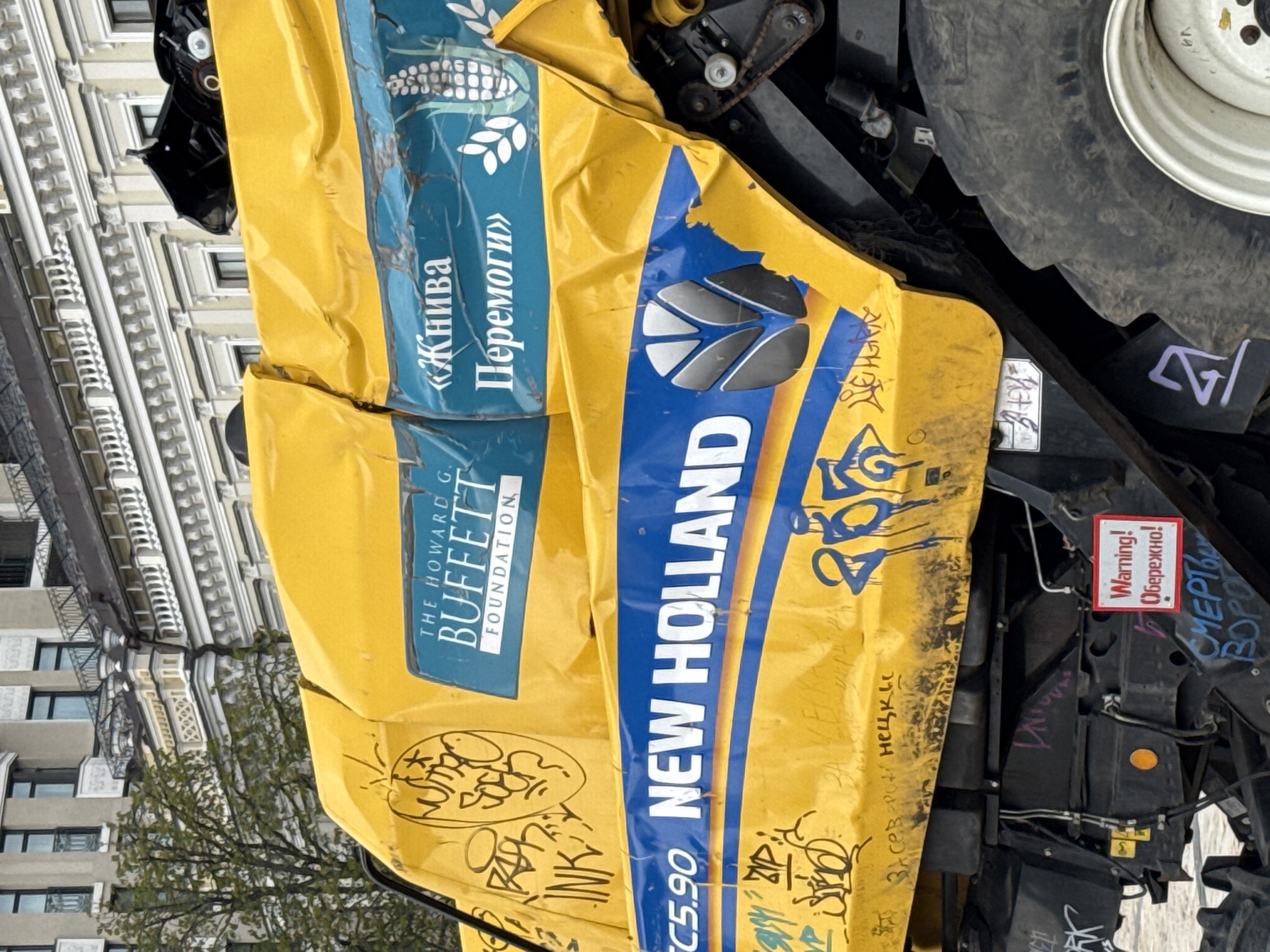 A yellow and blue New Holland TC5.90 combine harvester, visibly damaged and tagged with graffiti, sits in an urban environment. Markings include "THE HOWARD G. BUFFETT FOUNDATION" and the Ukrainian phrase "Жнива Перемоги" ("Harvest of Victory"). A warning sign reads "Warning! Обережно!" ("Caution!"). The surrounding cityscape suggests a location in Kyiv, Ukraine. A building and trees are visible.