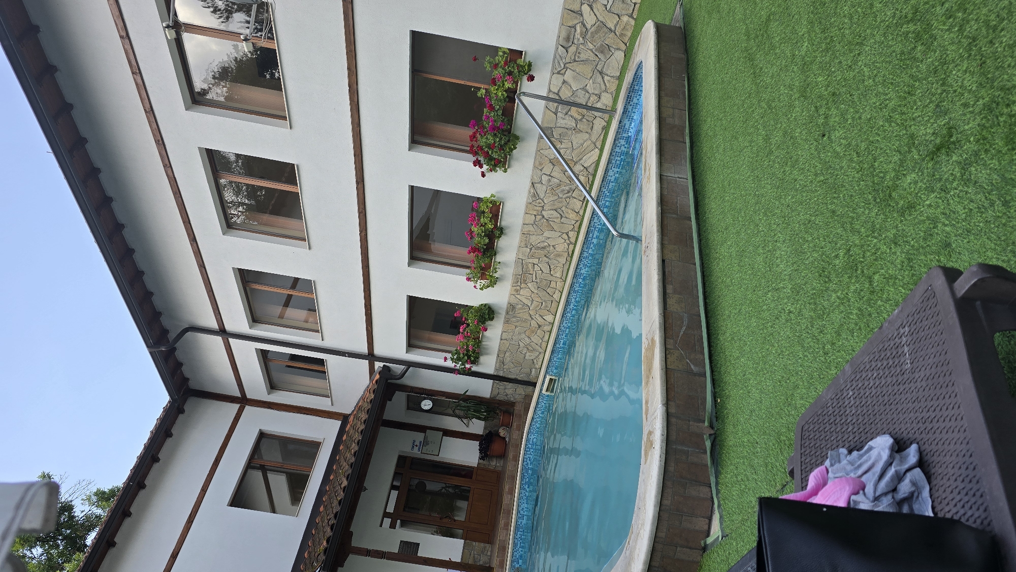 This is a photograph taken in the city of Strelcha, Bulgaria, depicting an outdoor scene with a swimming pool and a building. The building is painted white with dark trim and features several windows. Flower boxes filled with pink and red flowers are mounted beneath some of the windows. A stone facade is visible below these flower boxes. The swimming pool is turquoise blue, surrounded by a tiled edge and a stone border. A metal handrail extends into the pool. The ground surrounding the pool is covered with artificial green turf. In the foreground, a dark brown lounge chair is visible, with a black bag and some folded clothing on it. The sky is clear and blue.