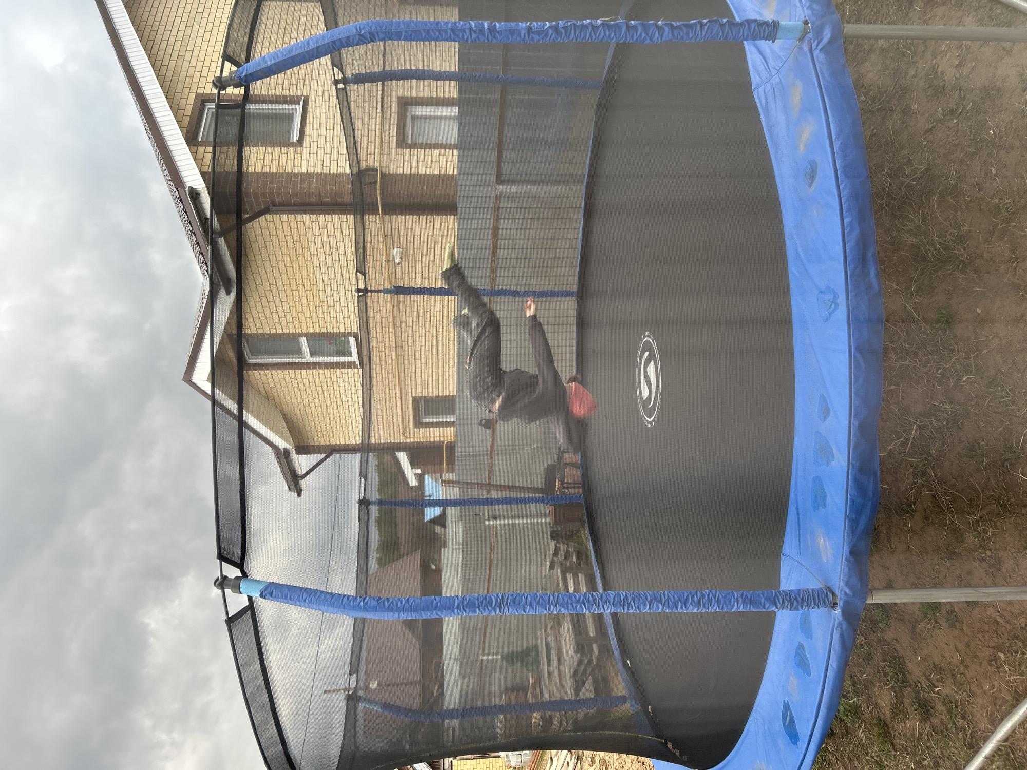 A color photograph taken in Chaikovsky, Russia, presents a close-up, angled view of a trampoline. A person wearing dark clothing and a red hat is in mid-air on the trampoline. The trampoline is surrounded by a safety net and a blue padded edge. The background shows a multi-story brick building with windows and a partial view of the overcast sky. Ground cover is visible to the right of the trampoline.