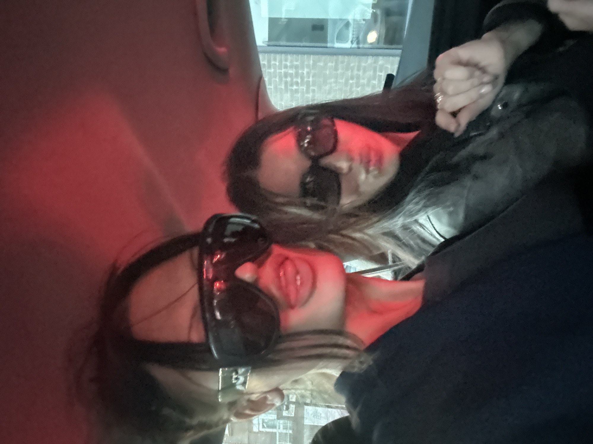 Two individuals are positioned inside a vehicle, possibly a car, given the interior lighting and visible elements of the car's interior. Both individuals are wearing sunglasses. The person in the foreground is sticking their tongue out. The interior of the vehicle is illuminated by a red light. A view outside the vehicle shows buildings typical of an urban environment. The specific location in London is not ascertainable from the provided image.