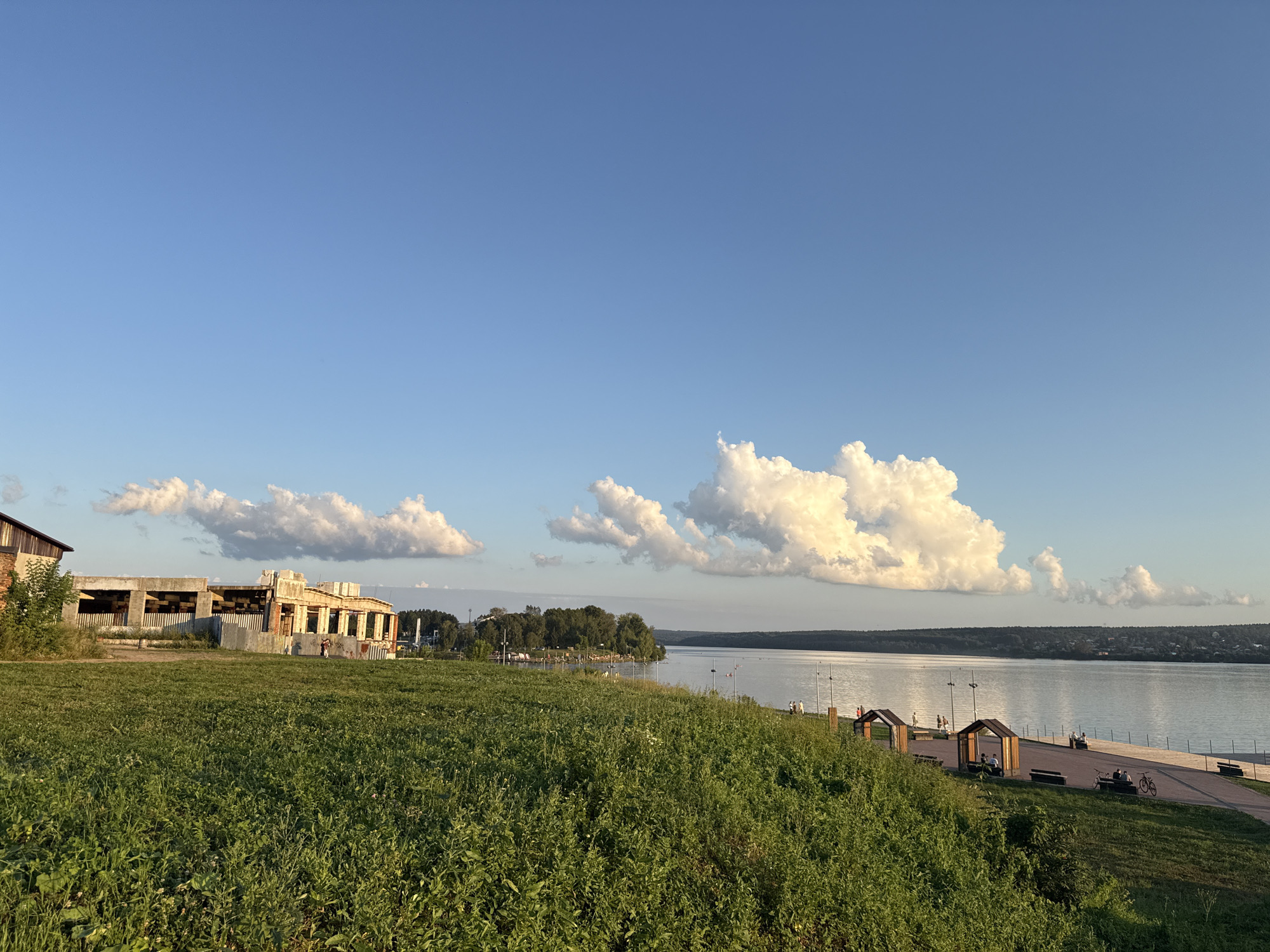 The image depicts a wide view of the Lysva River embankment in Lysva, Russia, under a clear blue sky with scattered white clouds. In the foreground, a lush green grassy hill slopes down towards the embankment. On the left side of the image, an unfinished building with exposed concrete pillars and a corrugated metal fence is visible. The embankment itself is paved and features several small, open-sided gazebos with wooden benches, some of which are occupied by people sitting or standing. A paved walkway runs alongside the river, and a bicycle is parked near one of the gazebos. Further along the embankment, there are more people strolling and enjoying the scenery. The Lysva River is wide and calm, reflecting the sky and the distant tree-lined shore on the opposite side. The overall impression is one of a pleasant summer day on a well-maintained urban waterfront.