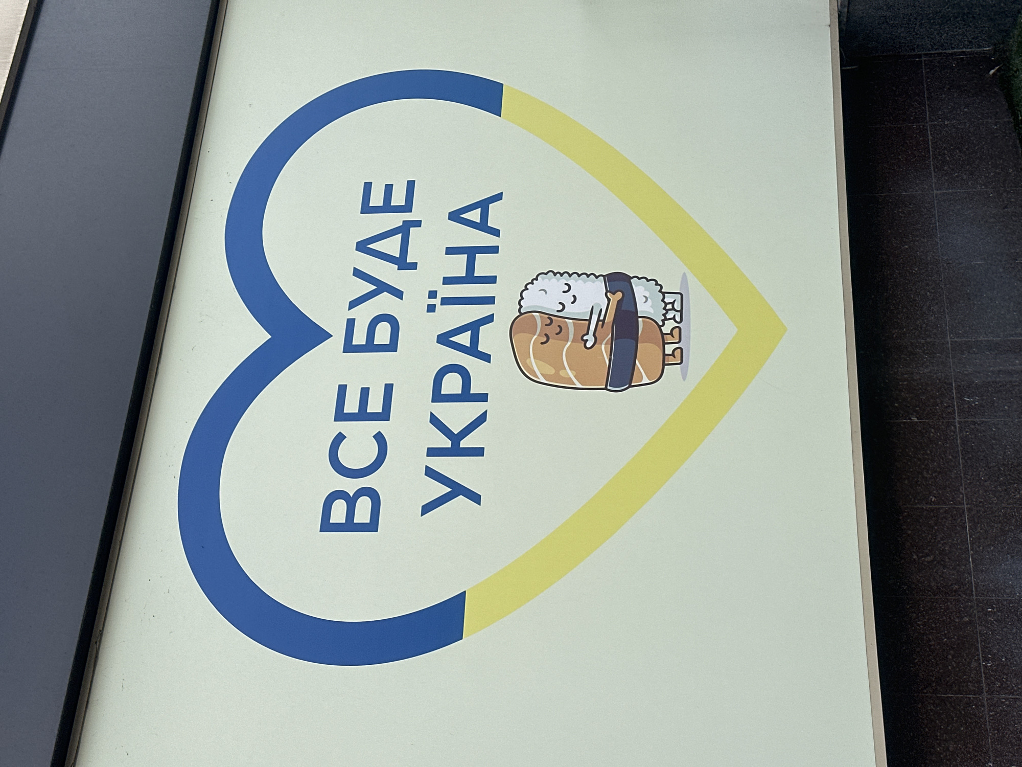 The image displays a vertically oriented sign or poster, likely situated in a public area in Kyiv, Ukraine. The main design element is a large heart symbol, distinctively colored blue on the left side and yellow on the right, mirroring the colors of the Ukrainian flag.
Within the heart, bold blue Cyrillic text reads "ВСЕ БУДЕ УКРАЇНА". This phrase translates to "Everything will be Ukraine" or "All will be Ukraine," a widely recognized patriotic slogan in the country, often interpreted as a statement of hope and resilience, implying that Ukraine will prevail.
To the right of the text, a charming cartoon character is depicted. It's a sleeping sushi roll, specifically a piece of salmon nigiri, characterized by its white rice base, orange-pink topping (salmon), and a dark band of seaweed. The sushi character has small, closed eyes, suggesting peaceful slumber, and appears to be snuggled under a small, dark object that resembles a pillow or blanket. Near its feet, a few small, white, cloud-like or ghost-like figures are visible.
The sign itself has a light, off-white background. On the left side of the image, a dark, vertical element, possibly part of a frame or adjacent structure, is visible. On the right, a dark, possibly tiled, ground or floor surface is partially seen. The overall impression is one of national pride, hope, and a gentle wish for peace, conveyed through a blend of national symbols and a comforting cartoon aesthetic.