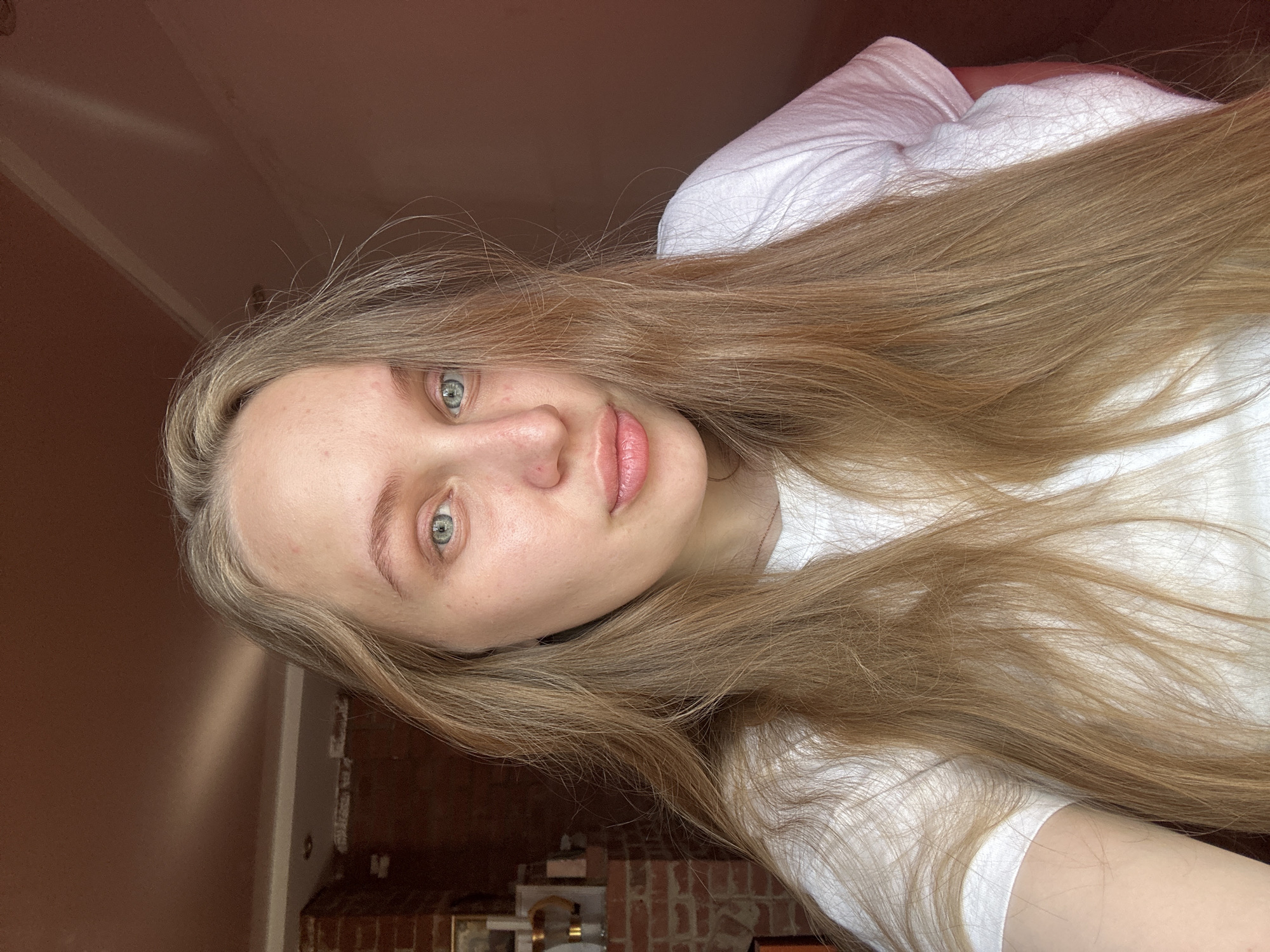 A young woman with long, blonde hair and blue eyes looks directly at the camera. She is wearing a white t-shirt. Her face is slightly turned to the left, and her lips are slightly parted. The background shows a brick wall and a ceiling with a diagonal shadow. The scene appears to be indoors. The image was taken in Chaikovsky, Russia.