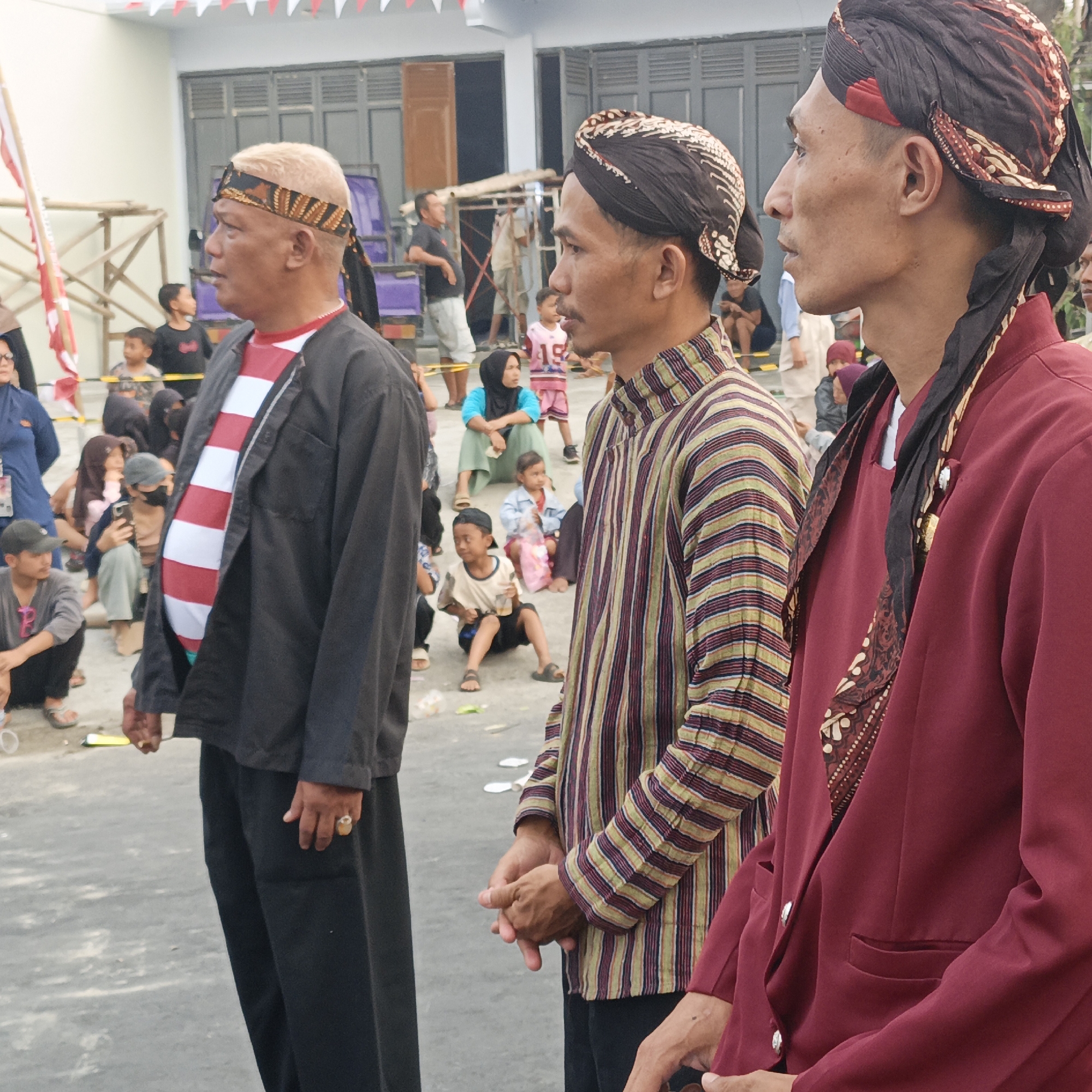 Three men, dressed in traditional Javanese attire, stand in an outdoor setting. The man on the left wears a black jacket over a red and white striped shirt and black pants, with a patterned headcloth. The middle man is dressed in a striped shirt and a dark patterned headcloth. The man on the right wears a maroon jacket with a patterned cloth accessory and a dark patterned headcloth. Several other people, including children and adults, are visible in the background, some seated. The ground is paved, and there are elements of event decorations such as flags. The overall impression is that of a cultural gathering or celebration. The location is specified as Randudongkal, Indonesia.