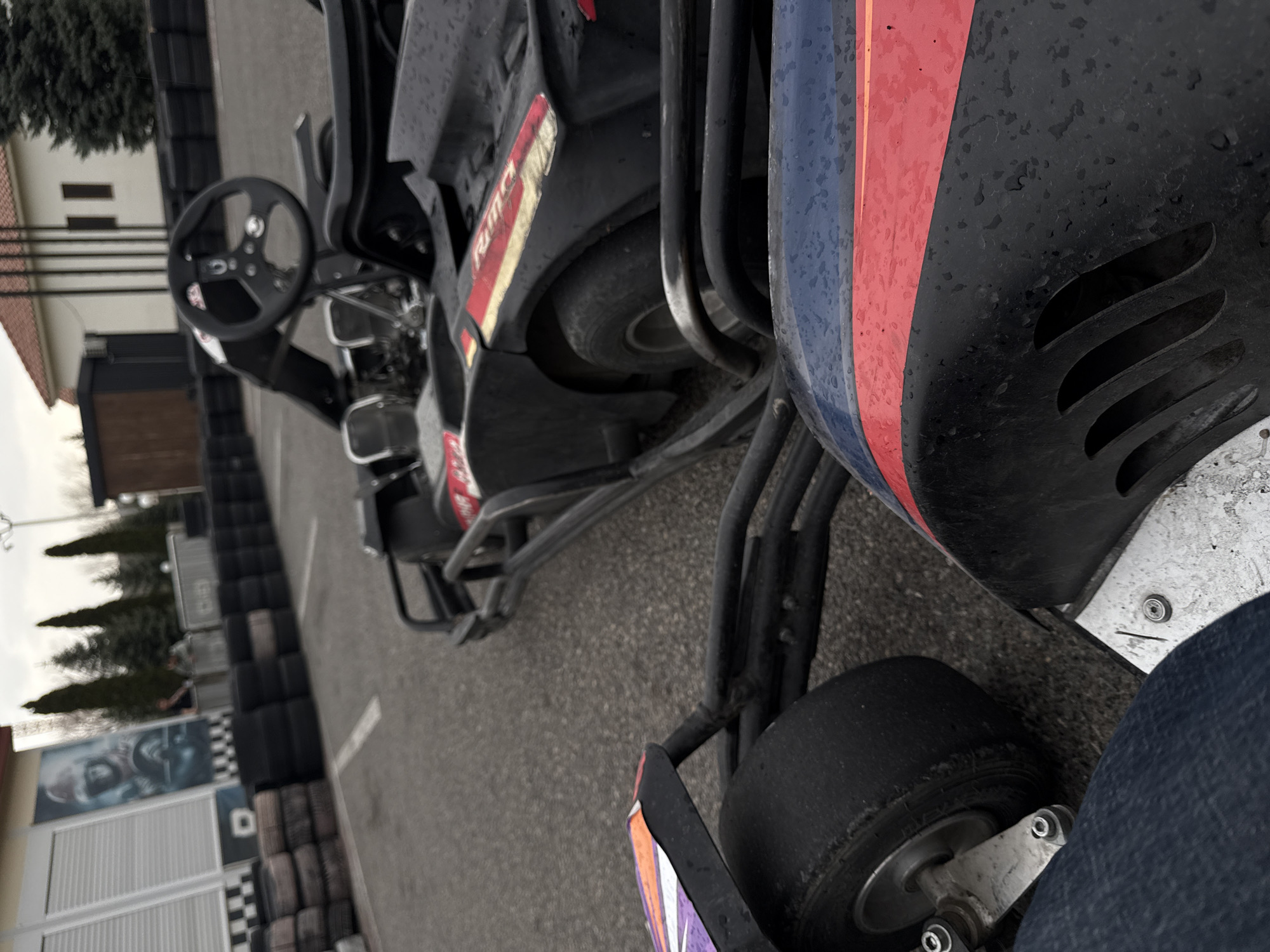 The image captures an outdoor go-karting track, viewed from a low angle. The sky is overcast, and water droplets are visible on the surfaces of the go-karts, indicating recent rain or damp conditions.
In the immediate foreground, part of a go-kart is visible, featuring a black, blue, and red fairing, along with its front right tire. A person's leg, clad in blue jeans, is also visible, suggesting they are seated in or near the go-kart.
Further into the scene, another go-kart is parked, predominantly black with red and white striped accents. Its steering wheel, chassis, and a rear tire are clearly visible. On the side of this go-kart, a red and white stripe contains partially visible text, which appears to include the letters "RMC".
The track surface is asphalt. Along the left edge of the track, numerous tires are stacked high, forming barriers. In the background, beyond the track, there are several buildings and trees. One building features a red-tiled roof, while another has roller shutters and a large poster displaying a close-up of a person's face. The letter "D" is also visible on this building.
The scene depicts a go-karting facility, likely during a break in activity or just before/after a session, in Yerevan, Armenia, under humid or wet weather conditions.