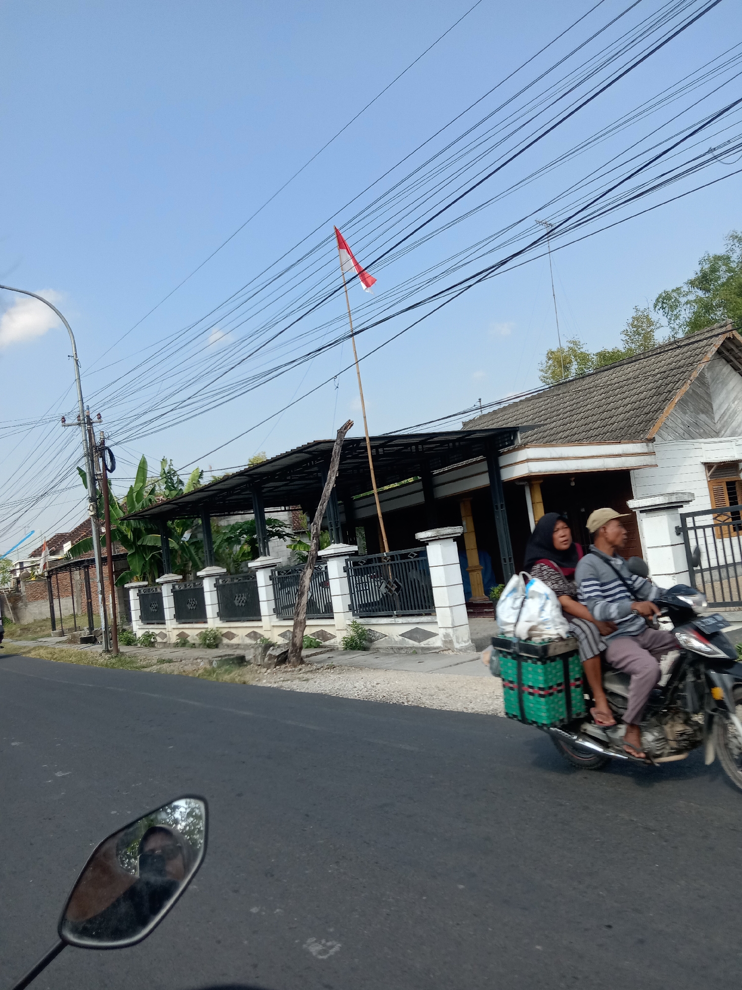 The image captures a street scene in Balongrejo, Indonesia, under a clear blue sky. In the foreground, a motorcycle is driven by a person wearing a light-colored cap and a striped long-sleeved shirt. A woman wearing a dark hijab and a patterned shirt is seated behind the driver. They are carrying two green plastic crates filled with white bags on the back of the motorcycle. To the left of the motorcycle, a motorcycle's rearview mirror reflects the face of the photographer. The background shows a white house with a tiled roof, a black fence with ornate white pillars, and lush green banana trees. A red and white Indonesian flag is visible on a bamboo pole above the house. Electrical wires crisscross the sky above the scene, supported by a utility pole on the left side of the image. The road is paved and appears to be asphalt.
