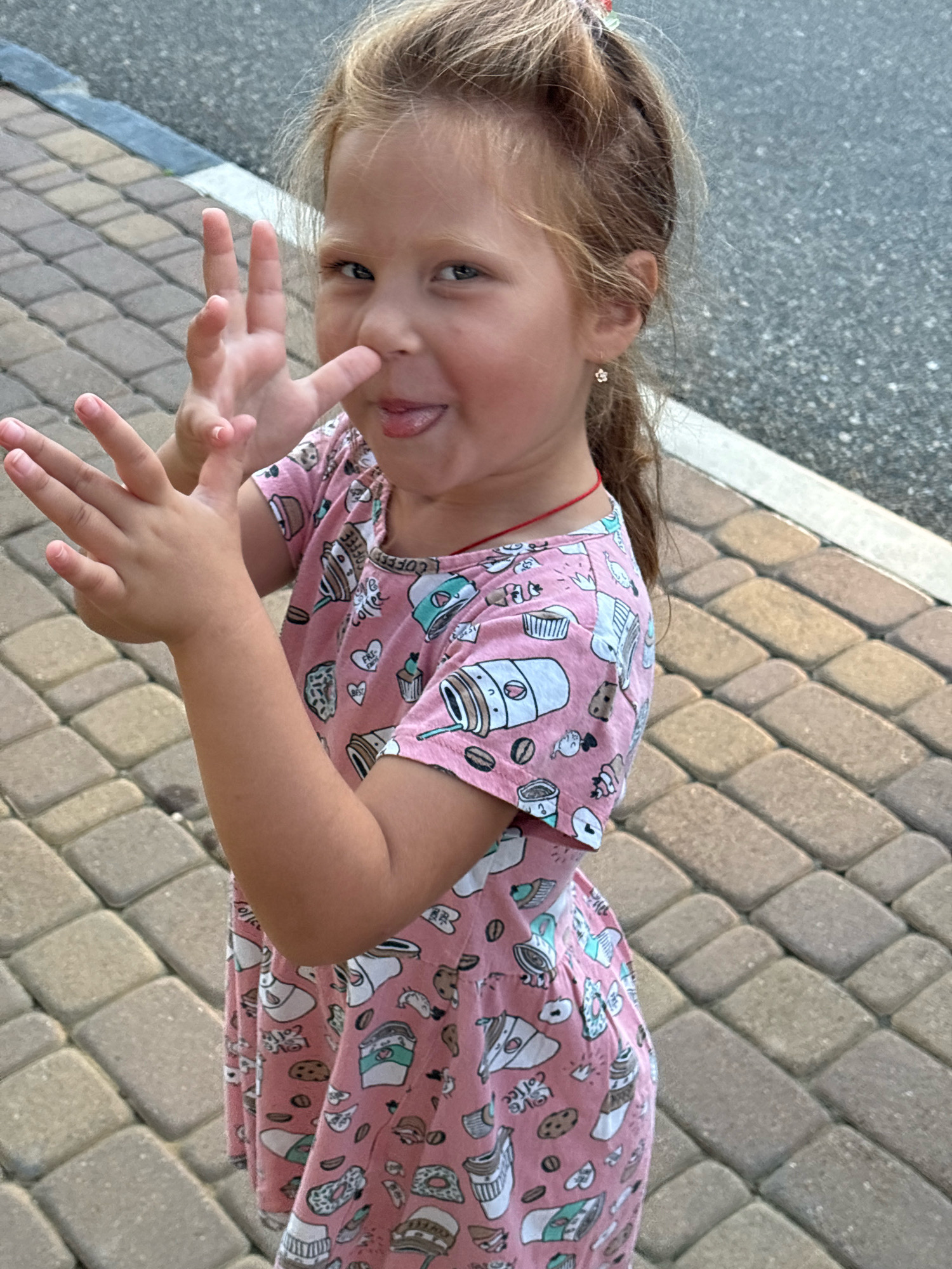 A young girl with blonde hair pulled back in a ponytail and a pink dress with a coffee-themed pattern stands on a patterned walkway, possibly in Arkhipo-Osipovka, Russia. She is sticking her tongue out and making a peace sign with her left hand, while her right hand is raised. The background shows a darker, rough surface, likely asphalt.