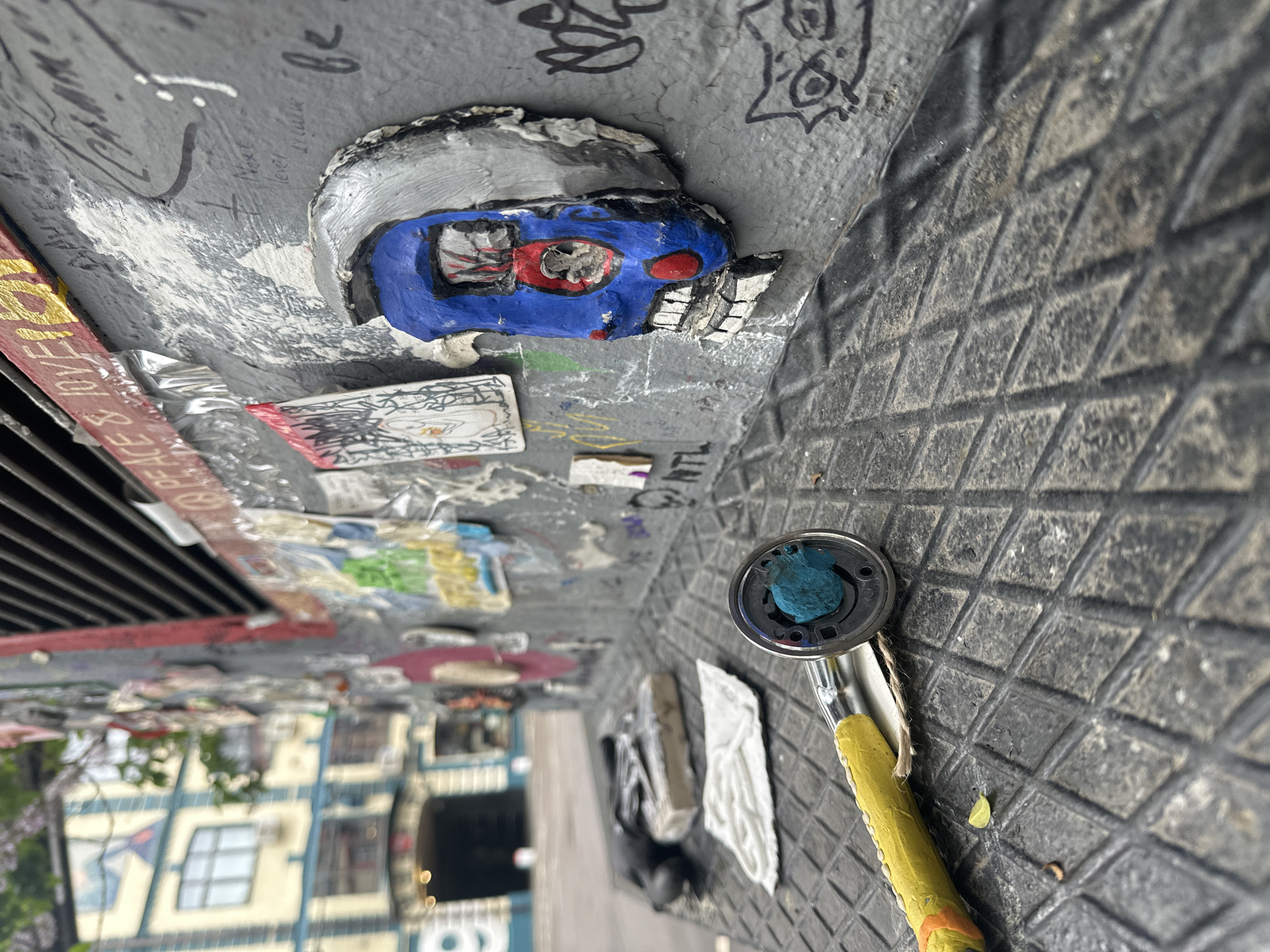 The image shows a street-level view looking upwards towards a metal surface covered in graffiti and street art. The art includes a three-dimensional blue and silver mask with exposed teeth, stickers, and various painted tags. Next to the mask, there's a metal grate above the street. Below the metal surface, there is a street scene, including parked cars and buildings with visible windows, and a small tree. In the lower right corner, there is a painted metal showerhead. The location is Moscow, Russia.