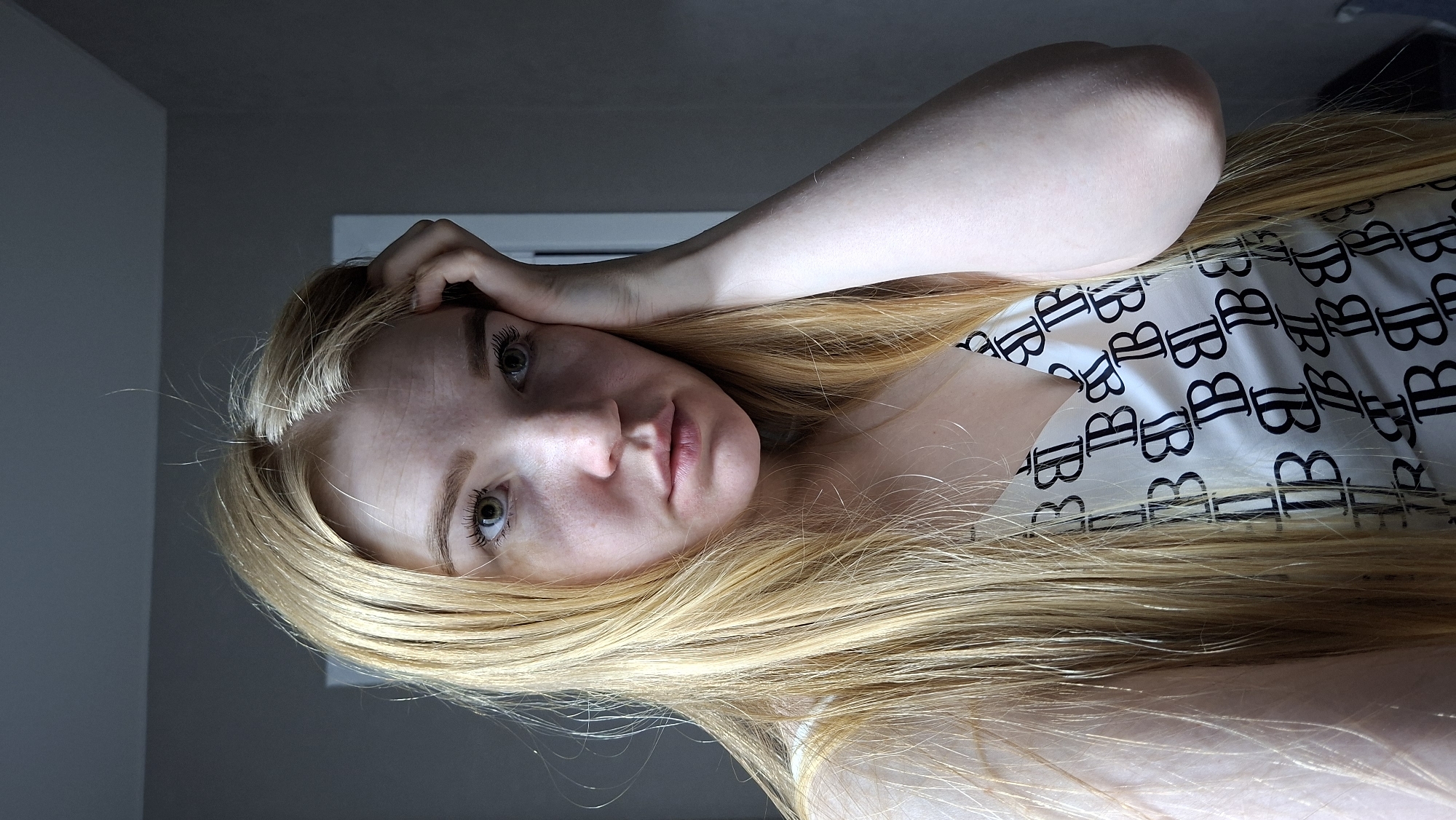 A fair-skinned woman with long, straight, blonde hair is depicted indoors. She is looking directly at the camera, with her left hand resting on her forehead. She is wearing a white top with a repeating black "B" pattern. The background consists of a gray wall and a window. The image's precise location in Chaikovsky, Russia, is undeterminable based on the visual content.