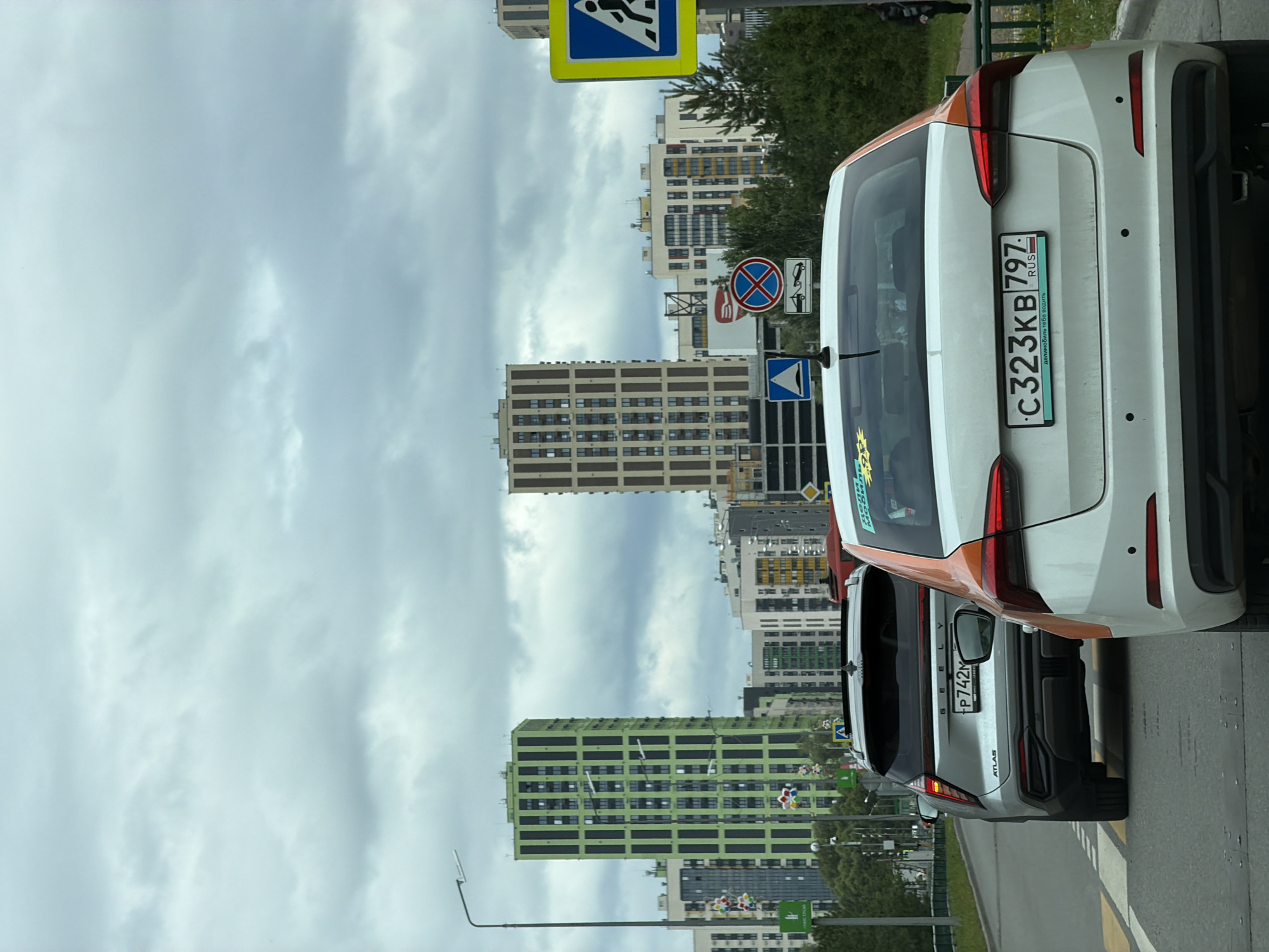 A vertical, eye-level, full shot captures a traffic scene with several cars and modern buildings under a cloudy sky in an urban environment. In the foreground, the rear of a white car with an orange stripe on the trunk is prominent, displaying a license plate with the characters "C323KB 797 RUS". To its left, a silver SUV, identified by the "GEELY ATLAS" badging on its rear and license plate "P742M", is also visible. Further down the road, a red vehicle is partially obscured.
The background features a cluster of tall, contemporary apartment buildings with distinct architectural styles. Two prominent buildings stand out: one is a tall, slender skyscraper with a vibrant green facade and dark vertical accents, and another is a taller, wider building with a beige facade and a pattern of recessed windows. Shorter buildings of varying colors and designs are also present, creating a dense urban landscape.
Streetlights, traffic signs, and trees line the road. A blue and yellow pedestrian crossing sign is visible on the right side of the frame, and another traffic sign with a red circle and a diagonal line, prohibiting something with an image of a tow truck below it, is situated further back. The sky is overcast with a light gray, textured cloud cover.