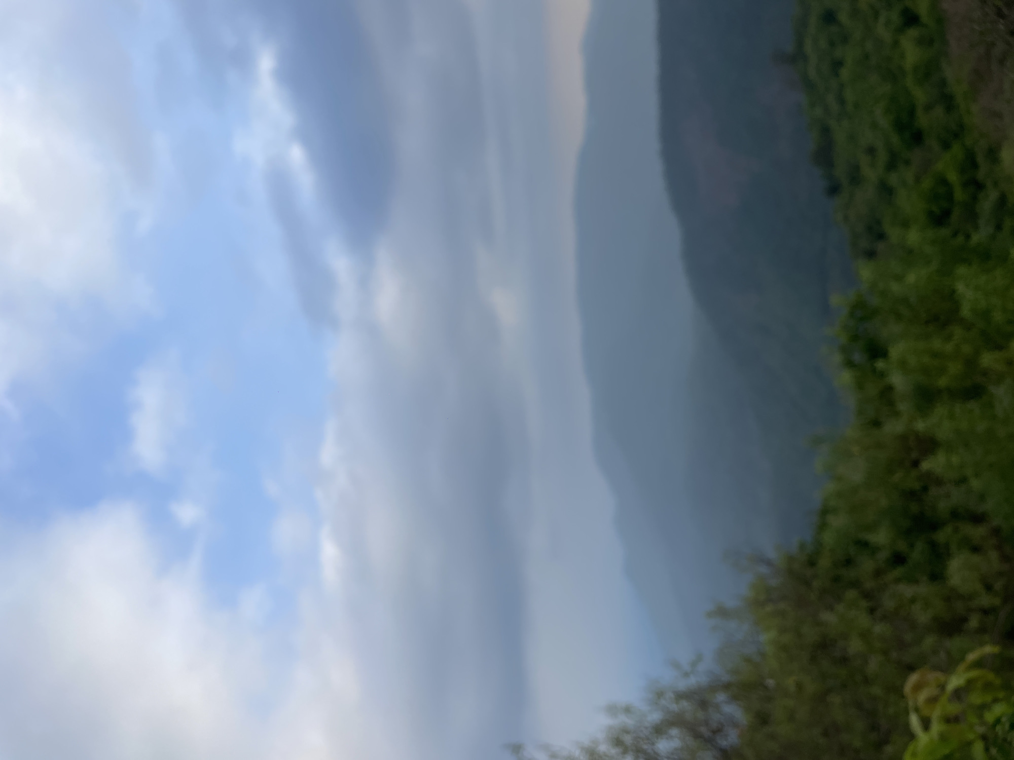 The image depicts a blurred landscape in Chorla, India. A cloudy sky fills the left side of the frame, transitioning to layered mountain ridges in the center, partially obscured by mist. Lush, green vegetation covers the right side of the image. The overall scene appears to be a natural, outdoor environment.