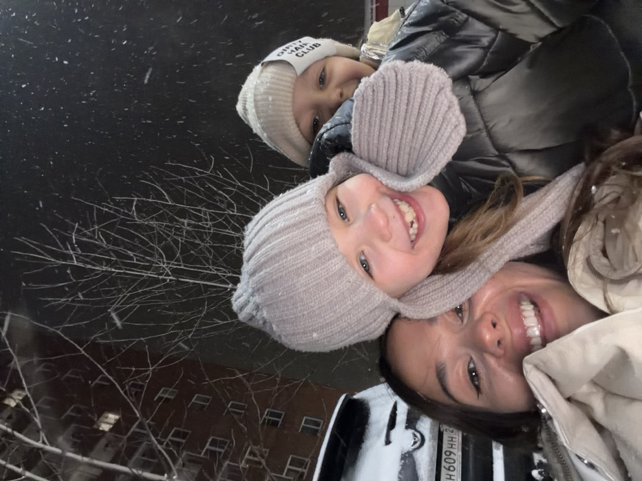 A nighttime scene depicts three individuals, an adult female and two children, in what appears to be a selfie-style photograph taken outdoors during a snowfall. All subjects are smiling.
The adult female, positioned in the lower left, displays a broad smile with visible teeth, and has long, dark hair. She wears a light-colored (possibly cream or beige) winter coat. A young girl, approximately 5-8 years old, is situated centrally, slightly behind and to the right of the adult. She has light brown hair, a wide smile with a visible gap from a missing front tooth, and wears a grey ribbed knit winter hat with ear flaps and a dark puffy winter jacket. A second, younger child is partially visible on the far right, mostly obscured by the central child. This child wears a cream-colored knit beanie with a white fabric tag bearing the text "DIRTY HAIR CLUB" and a shiny, possibly reflective, silver or light-colored winter jacket.
The background reveals an urban residential environment under active snowfall. To the left, a white or light-colored SUV or hatchback is parked, largely covered in snow. A Russian license plate, `H 609 HH 24`, is clearly visible on the vehicle's rear. Behind the vehicle and extending into the upper left, a dark, multi-story residential building with illuminated windows is discernible. Bare tree branches are silhouetted against the dark night sky, with numerous snowflakes visible in the air.
The location is identified as the city of Krasnoyarsk, Russia, based on the `24` region code present on the visible Russian license plate, which corresponds to Krasnoyarsk Krai. The scene is outdoors at night, characterized by falling snow and cold temperatures.