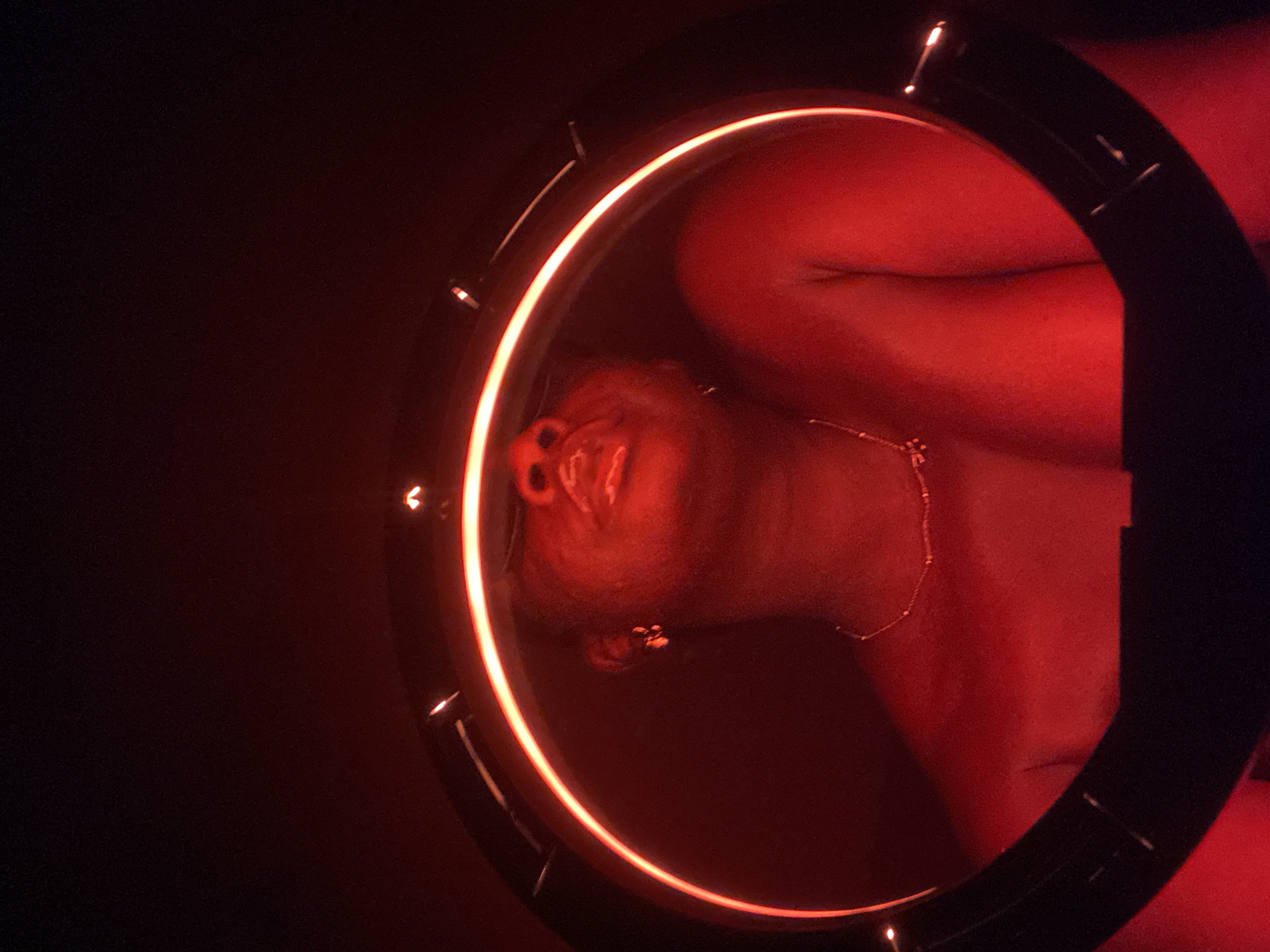 A person, illuminated by a red light ring, is oriented such that their head and upper torso are visible within the circle. The person is wearing a delicate necklace. The setting is dark and lacks discernible details to pinpoint a location in Maigizo, Nigeria.