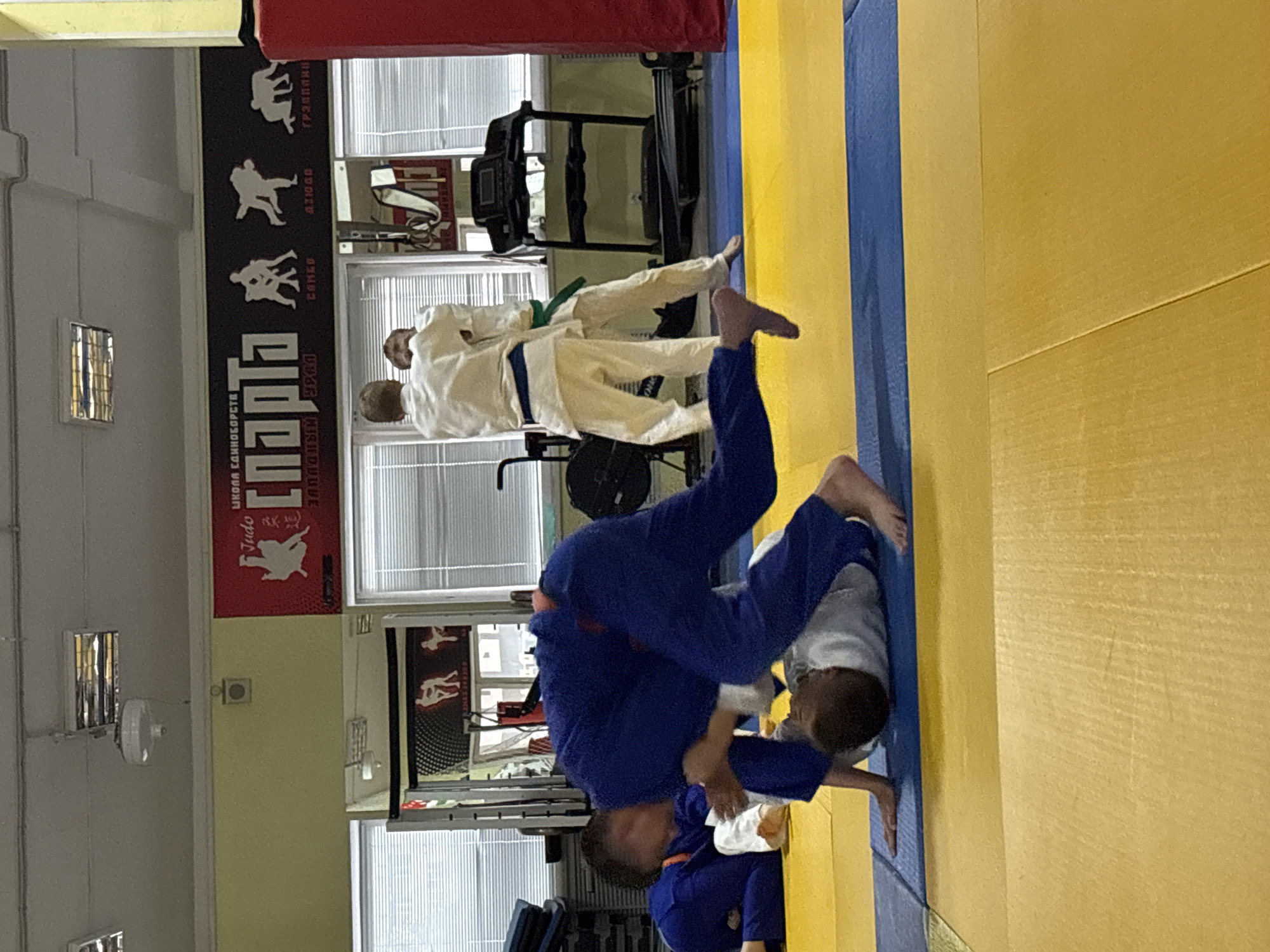 The image depicts a judo practice session. Two individuals in judogi are engaged in a grappling maneuver on a blue and yellow mat. One athlete is elevated, seemingly executing a throw, while the other is positioned below. Additional people in judogi stand in the background. The location is likely a judo training facility. The sign on the wall reads "Парта", indicating Judo club "Парта", city of Chaikovsky, Russia.