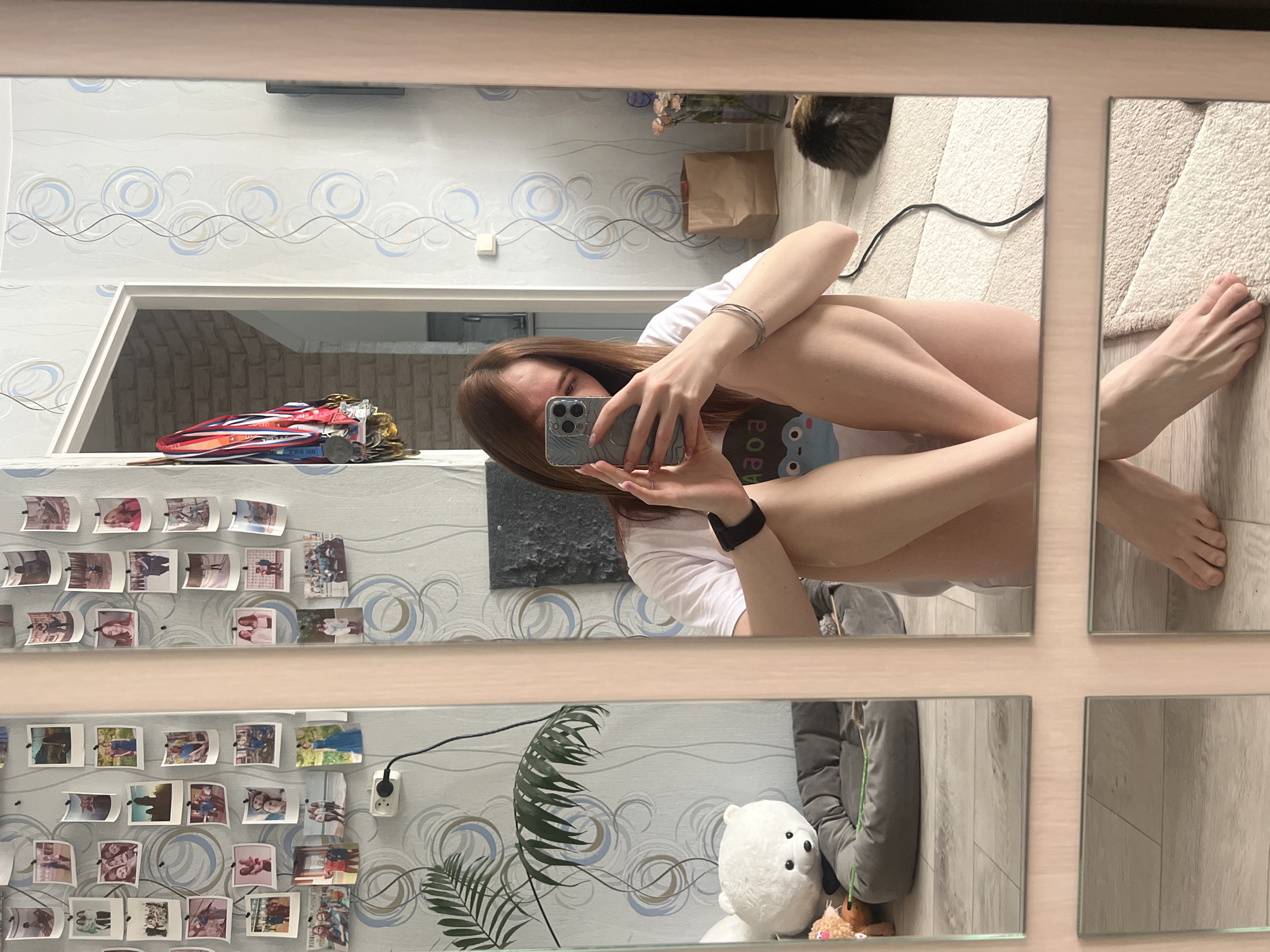 A selfie reflection in a multi-panel mirror. The person holding the phone has long brown hair, a white t-shirt with a blue graphic. The person is sitting with their knees up to their chest. The background includes a wall with photos, a doorway with what appear to be medals, a plant, a stuffed polar bear, and other household objects. There is a cat and shopping bag in the upper right corner of the image. The scene is located in an apartment. The image was potentially taken in Chaikovsky, Russia, as per the user's request.
