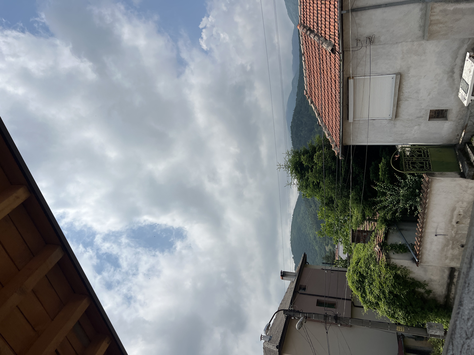 The image displays an outdoor, daytime scene in a mountainous, residential area. The perspective is slightly elevated, looking across buildings towards a distant range.
The upper portion of the frame is dominated by a partly cloudy sky, featuring a mix of light grey clouds and patches of clear blue sky. Power lines traverse the mid-ground horizontally.
On the right side of the image, the top edge of a wooden roof structure, with visible beams and dark trim, extends into the frame. On the left, a house with a red tiled roof and a light-colored, textured facade is prominent. This house features a window with a closed white rolling shutter, a small rectangular vent, and a decorative green metal gate below.
Further towards the bottom left, another building with a dark tiled roof and a chimney is visible, along with a utility pole equipped with two streetlights attached to its side. Lush green vegetation, including trees, bushes, and climbing plants covering parts of walls and distant roofs, is scattered throughout the scene. The background is filled with rolling mountains covered in dense green forest, appearing hazy.
The scene depicts a quiet, possibly rural or semi-rural setting. The light suggests it is daytime, with soft illumination indicating either an overcast sky or an early/late hour of the day. No people or active events are visible.
On a yellow and black sticker attached to the utility pole near the bottom left building, some faded and indistinct text, likely numbers or letters, is present, suggesting a utility or identification tag. The architecture and landscape are consistent with a village in a mountainous region like Topolovo, Bulgaria.