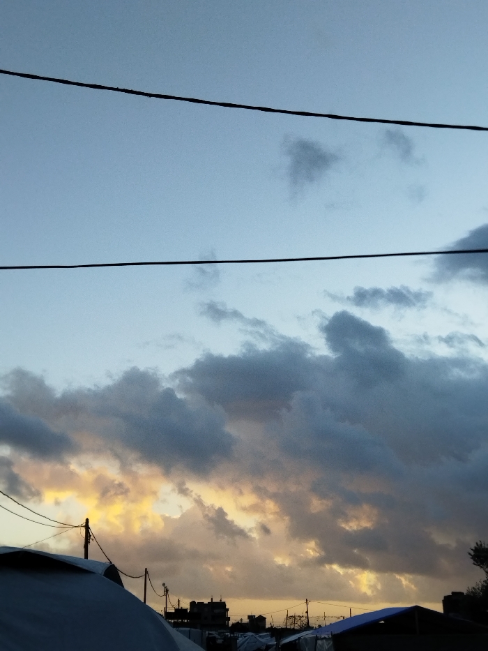 The image depicts a twilight scene over the city of Khan Yunis, Palestinian Territory. The sky occupies the majority of the frame, transitioning from a pale blue at the top to a dense formation of dark grey clouds in the middle and lower sections. Patches of intense yellow and orange light are visible between and behind these clouds, indicating either sunrise or sunset. Two prominent, dark horizontal utility lines traverse the upper portion of the sky. In the lower third of the image, the silhouettes of various structures are visible. On the far left, a large, light-colored, likely fabric-covered domed structure, possibly a tent, is partially visible. Multiple utility poles with entangled overhead wires are present across the mid-ground, standing against the illuminated sky. Behind these, indistinct silhouettes of multi-story buildings are discernable. Further to the right, more dark, low-lying structures, resembling tents or makeshift shelters, are visible, one featuring a distinct blue tarp. Sparse dark foliage is also present on the far right edge of the frame. No individuals are visible.