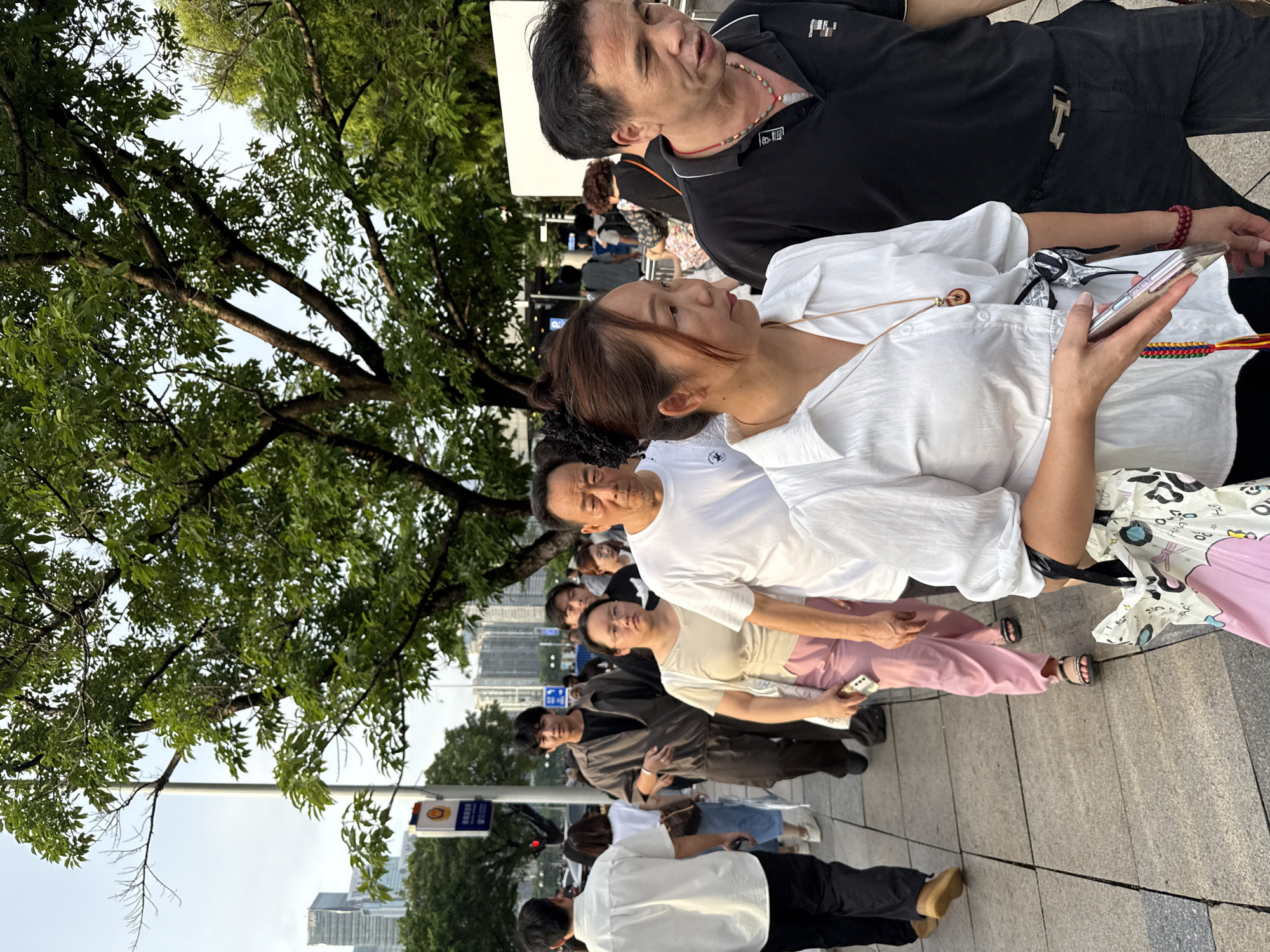 The image depicts a street scene in Haizhu District, China, with a group of people walking on a paved sidewalk lined with trees. In the foreground, a woman in a white blouse and a man in a black polo shirt are walking together. The woman is holding a mobile phone and has a necklace with a pendant. The man is wearing a necklace with red beads. Behind them, other individuals are walking in the same direction. A tall metal pole with signs is visible to the left, and in the background, modern buildings rise above the trees. The sky is overcast, suggesting it might be late afternoon or early evening.