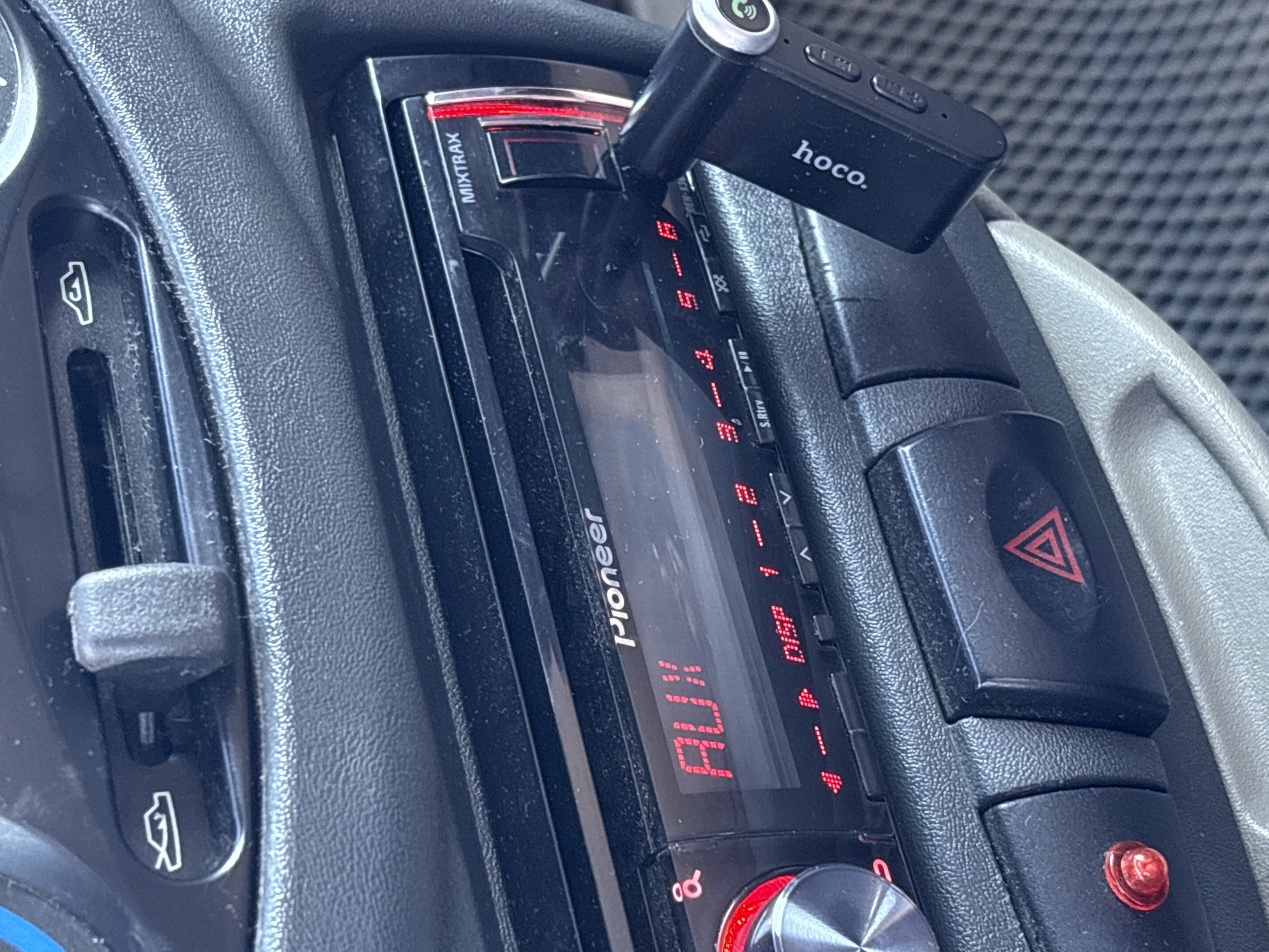 The image shows the interior of a vehicle, specifically the dashboard area. A Pioneer car stereo is prominently displayed, with the display illuminated showing "AUX". A black "hoco." branded bluetooth receiver is plugged into the stereo. To the right of the stereo are hazard light and other buttons. To the left, there appears to be an air vent control. The dashboard is textured black plastic. The environment appears to be the interior of a car. There is no evidence to confirm it is located in the city of Прикамский, Russia.