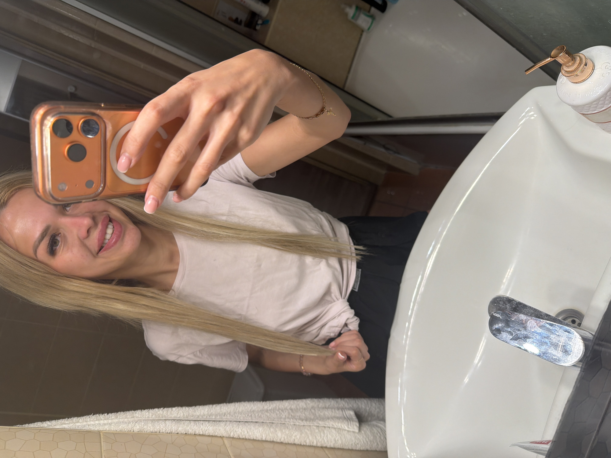 In a brightly lit bathroom in Kyiv, Ukraine, a woman with long blonde hair takes a mirror selfie. She is wearing a light pink t-shirt and black pants. Her left hand is holding an orange-cased smartphone, angled to capture her reflection. A white towel hangs on the left side of the frame, next to a beige tiled wall. In the foreground, a white sink with a chrome faucet is visible. A bottle with a rose gold dispenser sits on the right edge of the sink, reflecting the ambient light. The background shows a shower stall with glass doors and tiled walls.