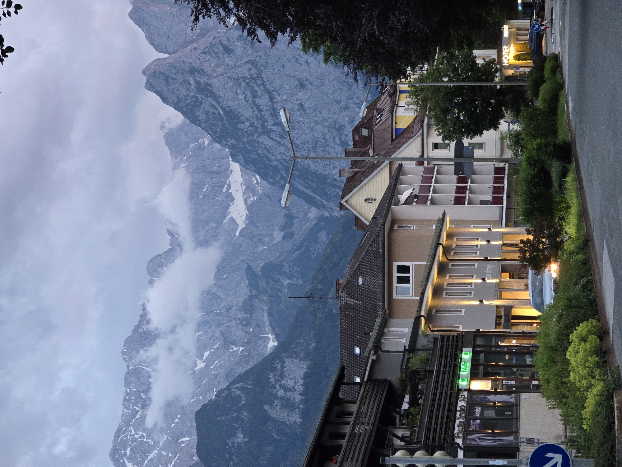 The image shows a street scene in Garmisch-Partenkirchen, Germany, with buildings on the left and a mountainous backdrop. In the foreground, a road curves to the right. Several buildings line the street, varying in architectural style. The building on the far left appears to be a multi-story establishment with balconies and shopfronts, one of which has illuminated green signage that reads "Friseur Salon Marrakech". Adjacent to it is another building with a light-colored facade and lit windows. Further down the street, more buildings with balconies and windows are visible.
Behind the buildings, a majestic mountain range dominates the background. The mountains are rugged and appear to be snow-capped in some areas, with clouds swirling around their peaks. The sky is overcast, contributing to a muted light in the scene.
In the middle ground, a street lamp stands tall, illuminating the area. A few trees are scattered along the street and in front of the buildings, adding greenery to the urban landscape. A car is visible on the road, and a traffic light with a blue directional arrow sign is also present. The overall impression is a quiet evening in a town nestled at the foot of impressive mountains.