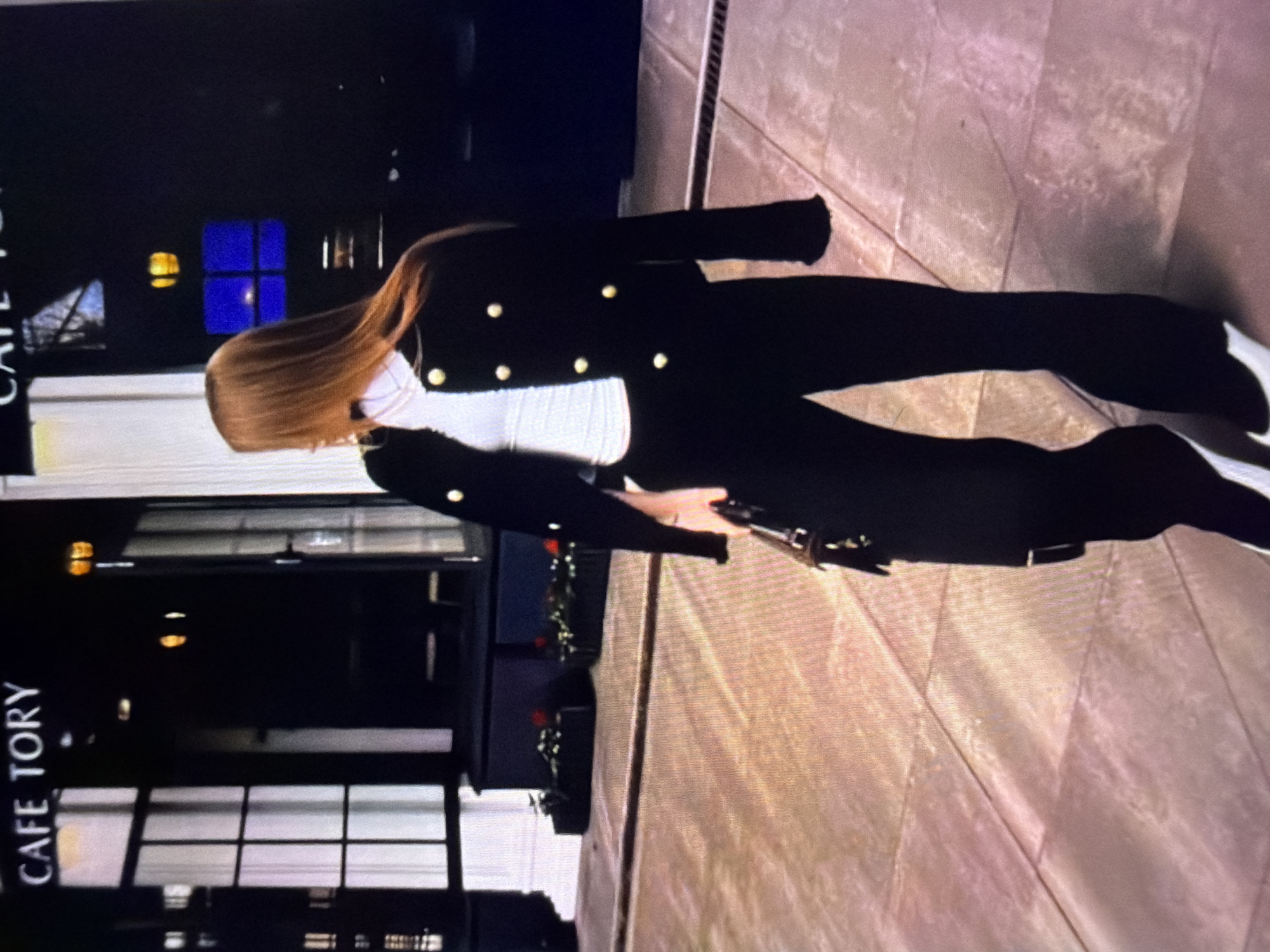 The image shows a person standing outdoors on a tiled floor, possibly in front of a building. The person is wearing a black jacket with gold buttons over a white top and black pants. Their hair is long and brown, and they are holding a silver object in their right hand. The background is dark, with some illuminated windows and a sign that reads "CAFE TORY" vertically. The overall lighting suggests it might be evening or night. The ground is paved with large, rectangular tiles in shades of beige and pink. The image is oriented vertically, with the person appearing to be upside down.