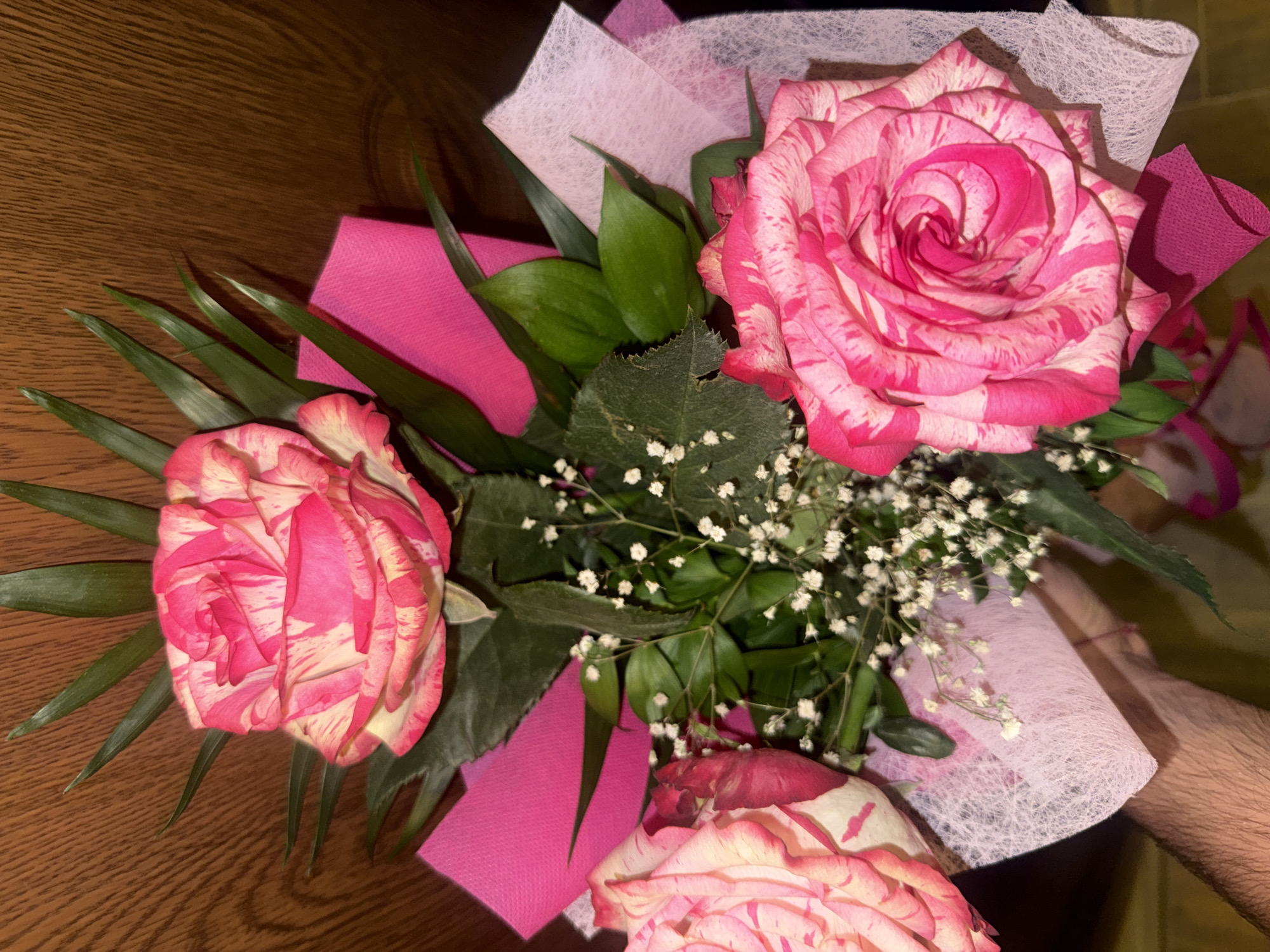 A bouquet of three pink and white striped roses, accented with small white flowers and green foliage, is presented against a wooden background. The bouquet is wrapped in pink and white tissue paper. The image appears to be taken indoors. No specific location in Varna, Bulgaria can be identified from the provided image.