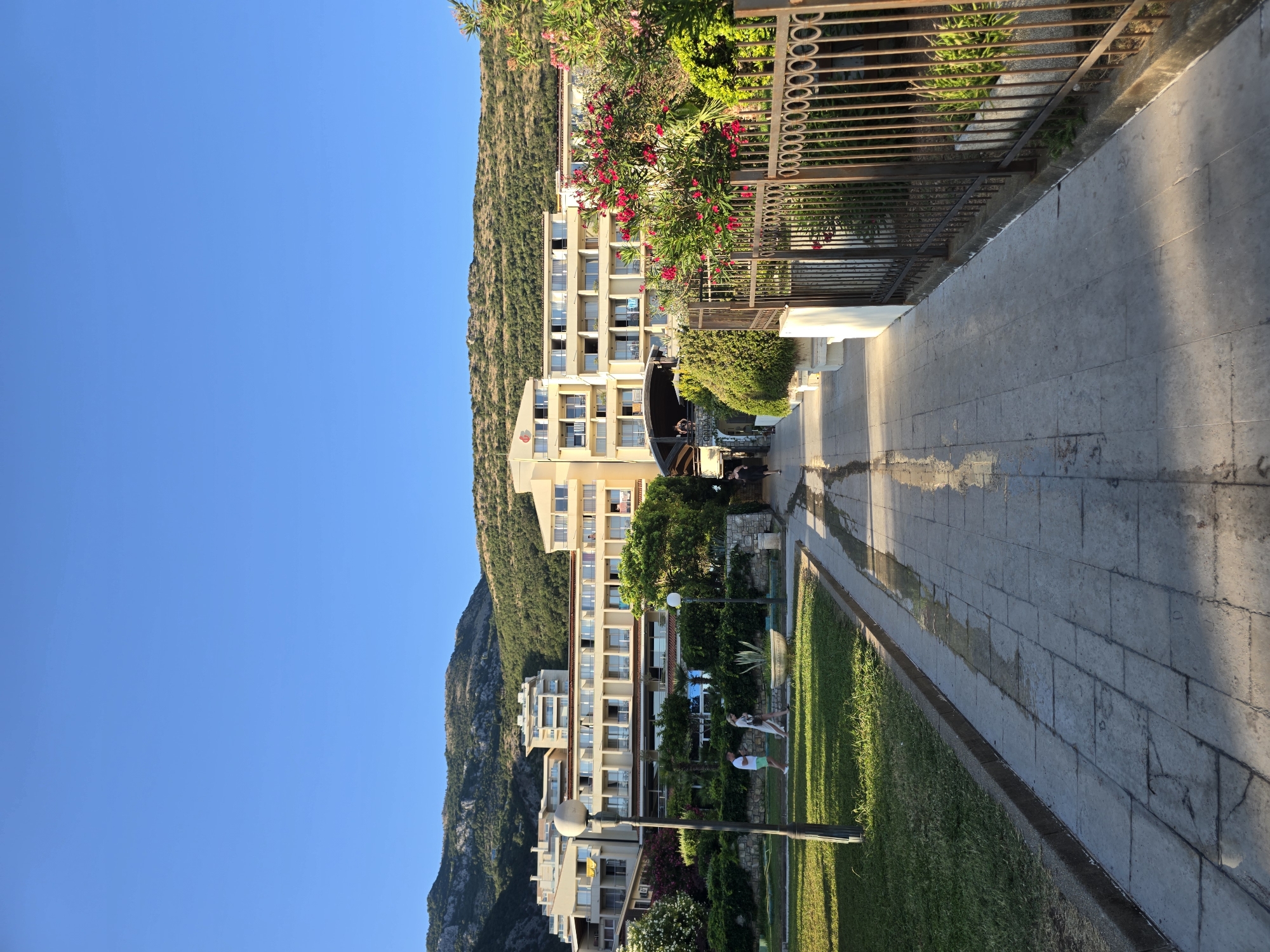 The image depicts a pathway leading towards a multi-story hotel complex situated at the base of a lush green, mountainous landscape. The sky is a clear, bright blue. The pathway is paved with light-colored square tiles and appears to be wet in places, reflecting the sunlight. On the left side of the pathway, there is a well-maintained lawn with a lamppost and two individuals walking away from the camera. A dark metal fence runs along the right side of the pathway, bordering a garden area with blooming pink flowers and dense green foliage. The hotel itself is a cream-colored building with multiple balconies and windows. The scene is consistent with the city of Petrovac centar, Montenegro.