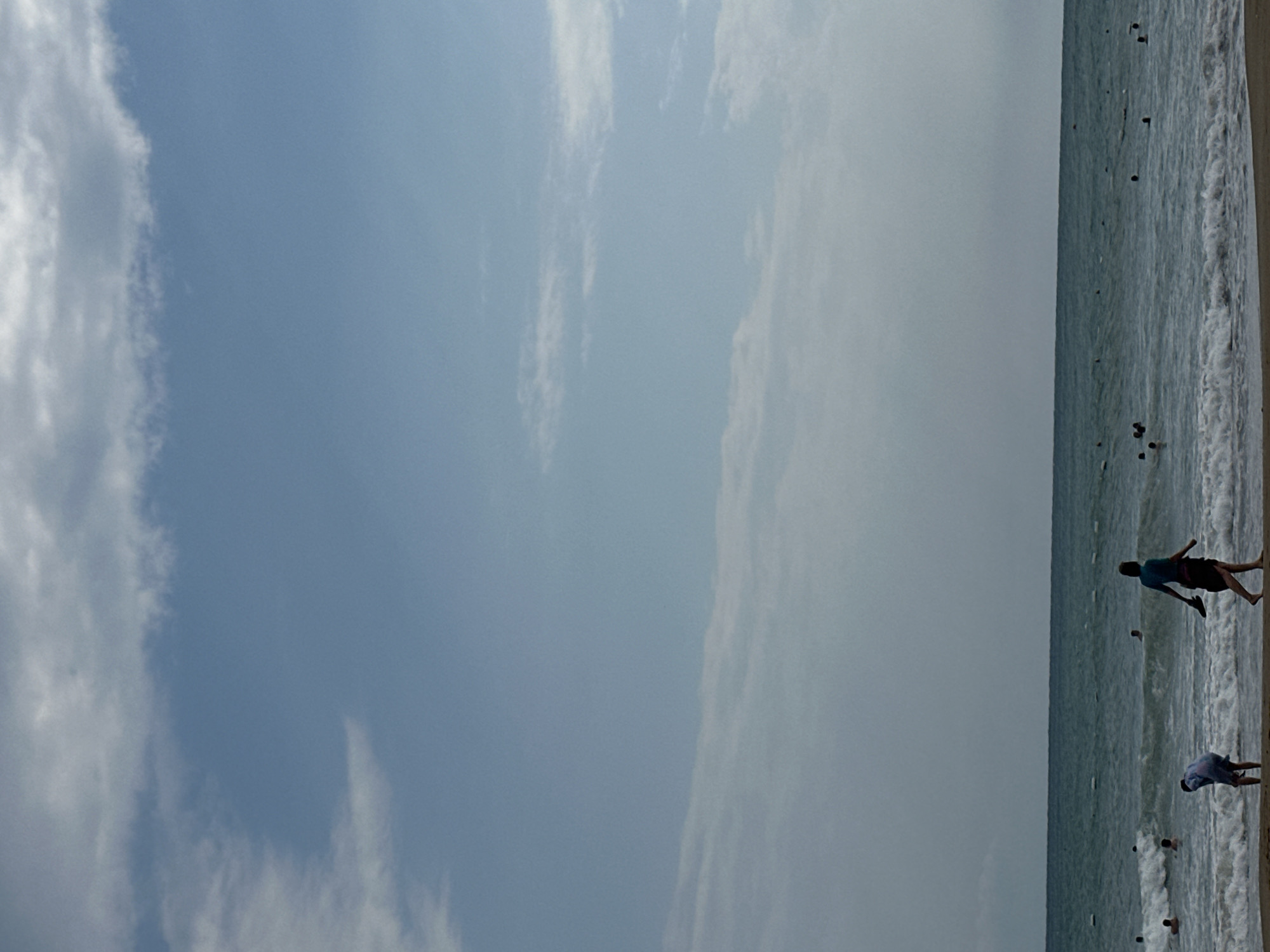 This is a wide shot of a beach in Manavgat, Turkey, on a day with a mostly blue sky scattered with white clouds. The ocean is visible in the mid-ground, with moderate waves breaking on the shore. A sandy beach occupies the foreground. Several people are present in the scene: two individuals are on the beach in the foreground, one bending over and the other walking away from the camera. Numerous other people are in the water, some swimming and others appearing to be further out in the sea. The overall atmosphere is that of a typical summer day at the beach.