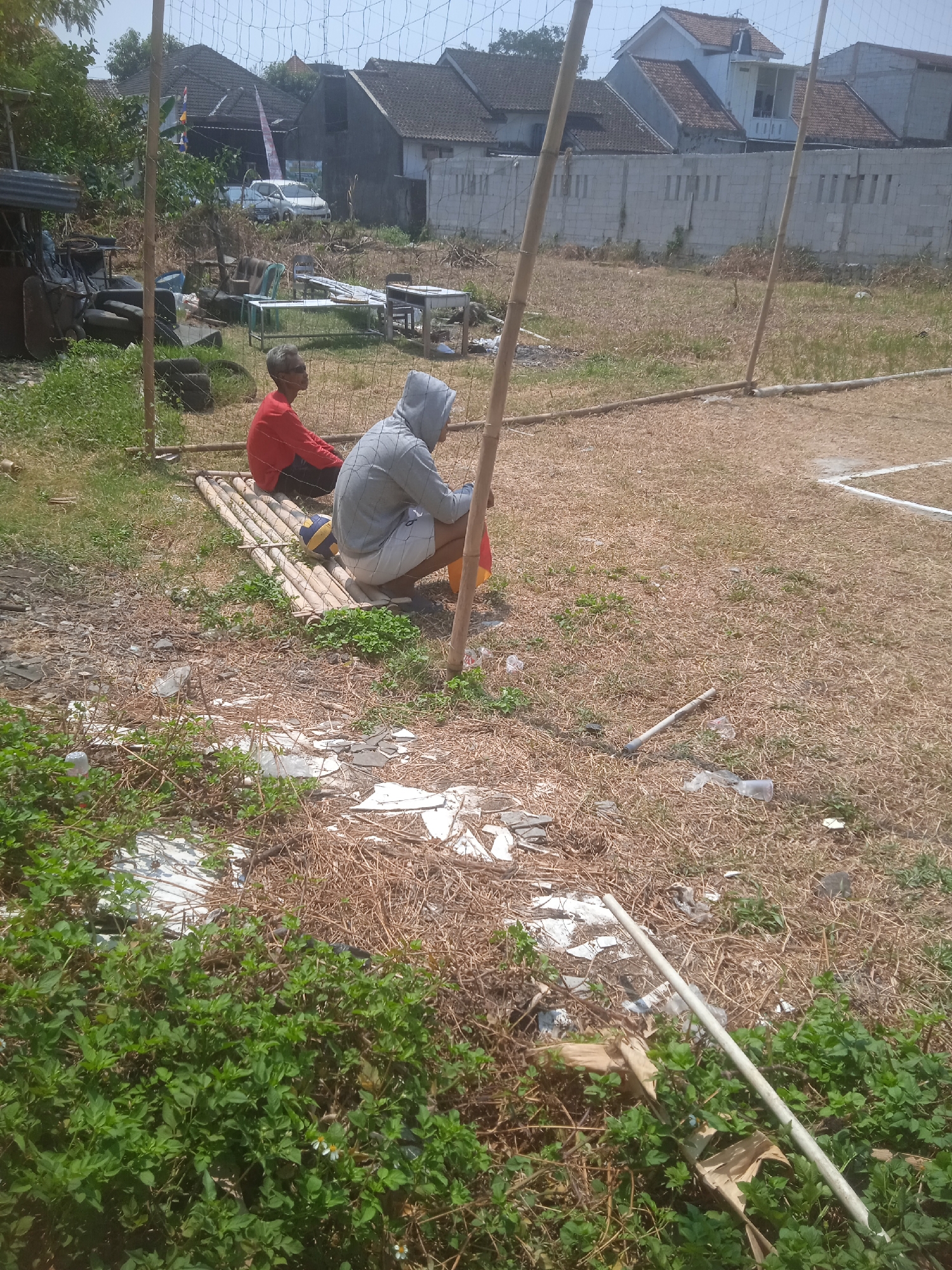 In Klodran, Indonesia, two individuals are observed seated on a makeshift raft constructed from bamboo poles. The person on the left, wearing a red long-sleeved shirt, faces away from the camera. Beside them, a person in a gray hoodie and white shorts sits, looking towards the right. A volleyball is positioned between their legs. The immediate surroundings depict a dry, overgrown field littered with debris, including plastic bottles, paper scraps, and broken white tiles. In the background, a white concrete wall stretches horizontally, beyond which are houses with tiled roofs and a clear sky. A net, possibly for a sports court, is visible in the foreground, supported by bamboo poles. The scene suggests an outdoor, possibly abandoned or underutilized, recreational area.