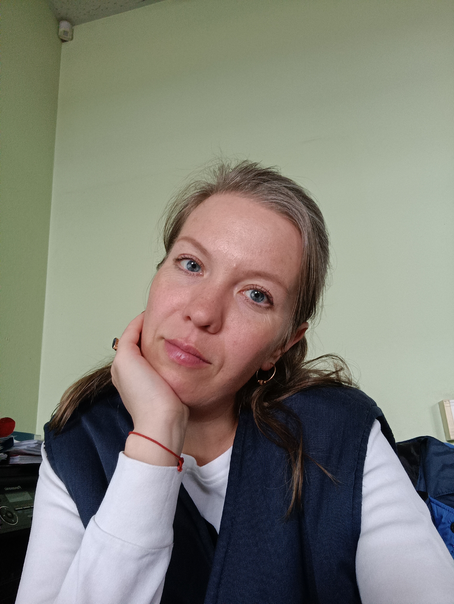 A woman is shown in a close-up, head-and-shoulders shot. She has light skin, blue eyes, and light brown hair with some graying at the roots. She is wearing a white long-sleeved shirt with a navy blue vest. She has a red string bracelet on her left wrist and gold hoop earrings. Her left hand is supporting her chin. The background is a light green wall, and a white security camera is mounted in the upper left corner. A stereo is visible behind her.
There is no evidence in the image to suggest that this photo was taken in the city of Chaikovsky, Russia.