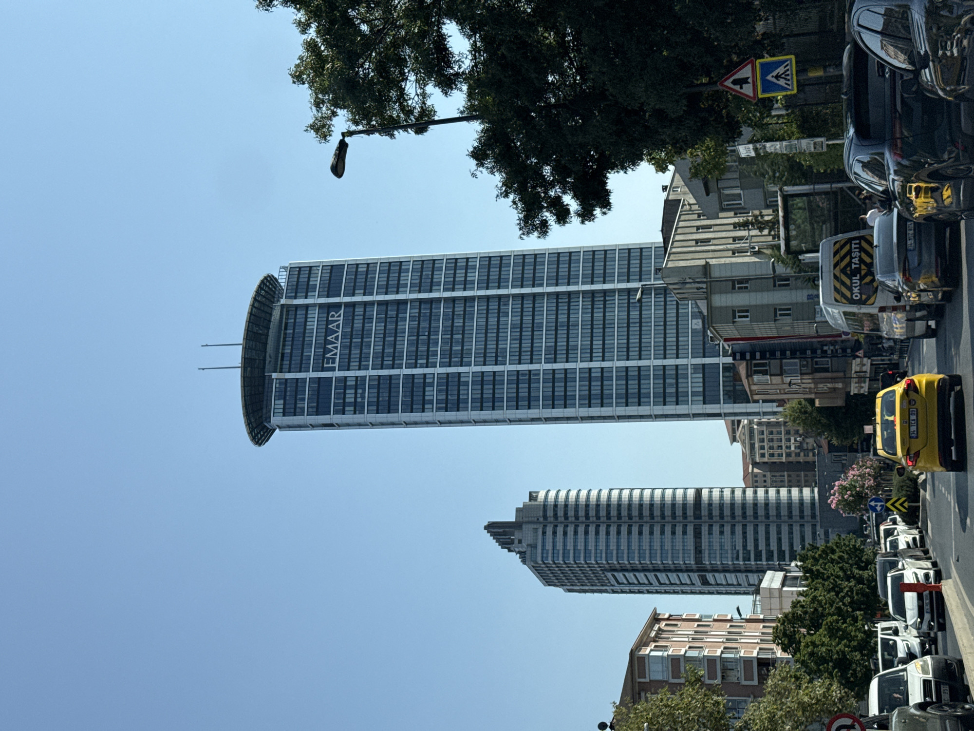 This is a full-body, eye-level, medium shot captured on a sunny day of a street in Üsküdar, Turkey, featuring traffic and modern skyscrapers. The foreground shows a street with several vehicles, including a yellow taxi in the right lane, a dark SUV behind it, and other cars and vans in the left lane. A white van with the text "OKUL TAŞITI" (school transport in Turkish) is visible. There are traffic signs, including a pedestrian crossing sign and a sign with a circular arrow indicating a no-entry zone or a one-way street. Trees line the right side of the street.
In the background, two prominent skyscrapers dominate the skyline. The taller one on the right is a sleek, modern building with a circular helipad on top and "EMAAR" clearly printed on its facade. To its left, another tall, modern skyscraper with a distinctive tiered top stands. The sky is clear and blue. The overall impression is a bustling urban environment with a blend of contemporary architecture and daily city life.