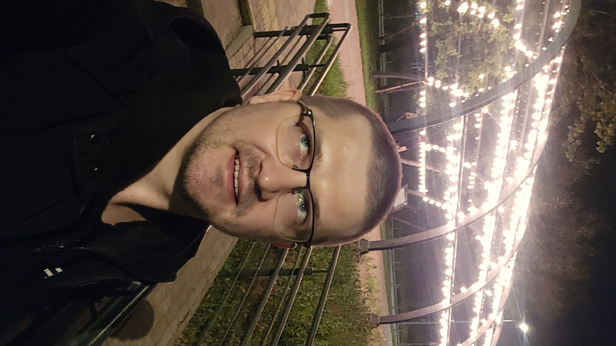 A man wearing glasses and a black hooded jacket is photographed from the chest up, looking towards the upper left of the frame. He has short hair and a light beard. Behind him, an arched structure is illuminated by numerous string lights. The background shows some greenery and a railing on the left. The scene appears to be outdoors at night. The location is the Hydropark in Kyiv, Ukraine, near the pedestrian bridge.