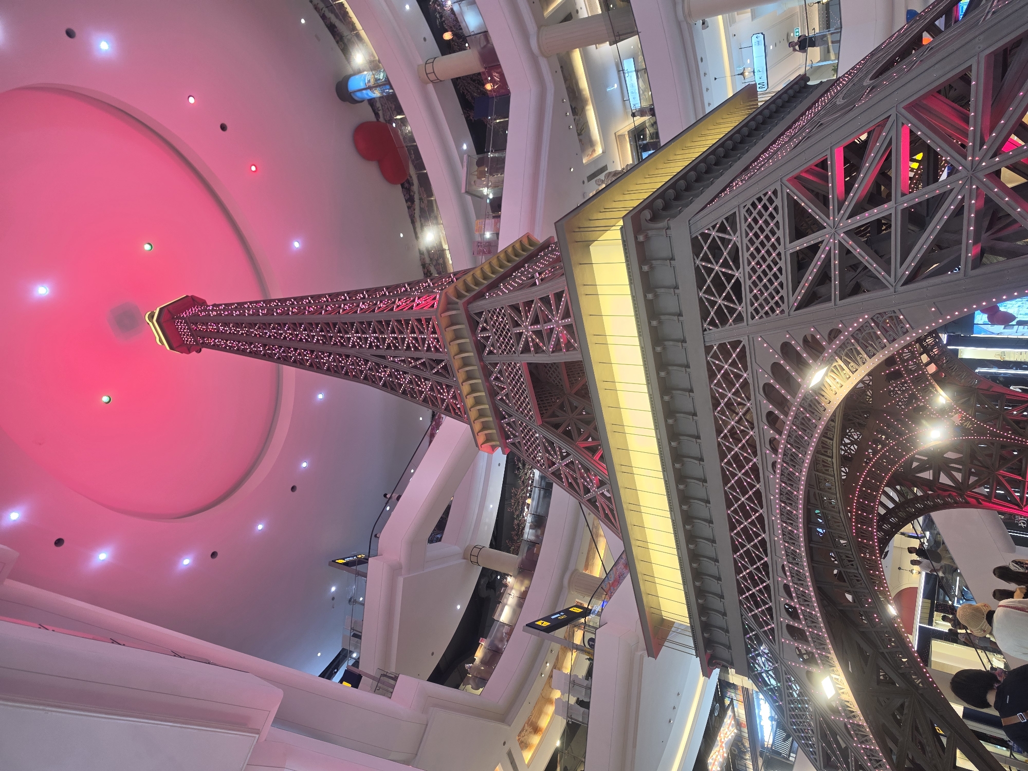 The image shows a low-angle view of a replica of the Eiffel Tower inside a shopping mall. The tower is made of steel and is lit with lights that alternate between pink and white. The tower is surrounded by shops and restaurants. The ceiling above the tower is round and lit with pink lights. People are standing around the base of the tower. The location is Pattaya City, Thailand.