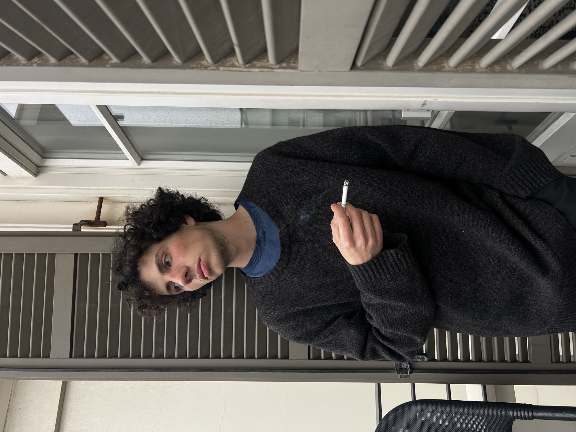 A young man with curly dark hair stands in front of a window and a metal railing, holding a lit cigarette. He wears a dark gray sweater over a blue shirt. The background includes a window with a white frame, closed white shutters, and a metal railing. The photo was likely taken in Tel Aviv, Israel. The man appears to be outside on a balcony or patio.