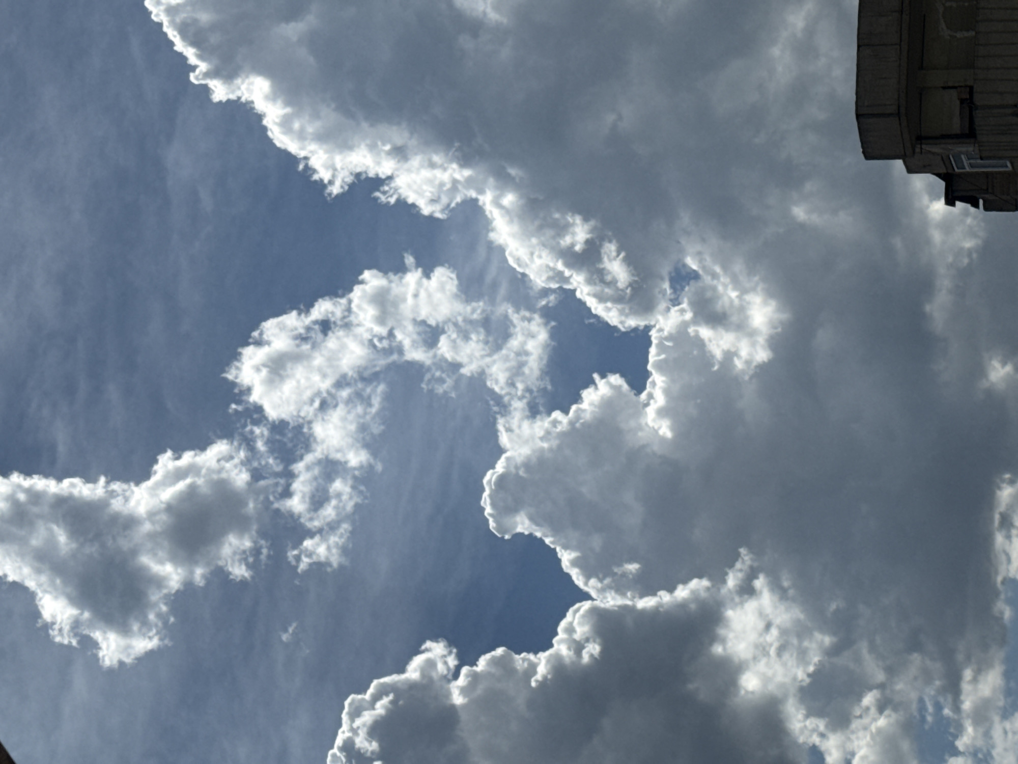 The image captures a dramatic sky with a mix of bright white and dark gray cumulus clouds against a clear blue sky. The clouds are voluminous and textured, with sunlight illuminating their edges and creating stark contrasts. In the lower right corner, a portion of a building with a flat roof and balconies is visible, suggesting an urban environment. The building appears to be made of concrete. The scene is likely an outdoor view from Abovyan, Armenia, looking upwards at the sky, possibly during a day with variable weather conditions. There are no people or animals visible in the image.