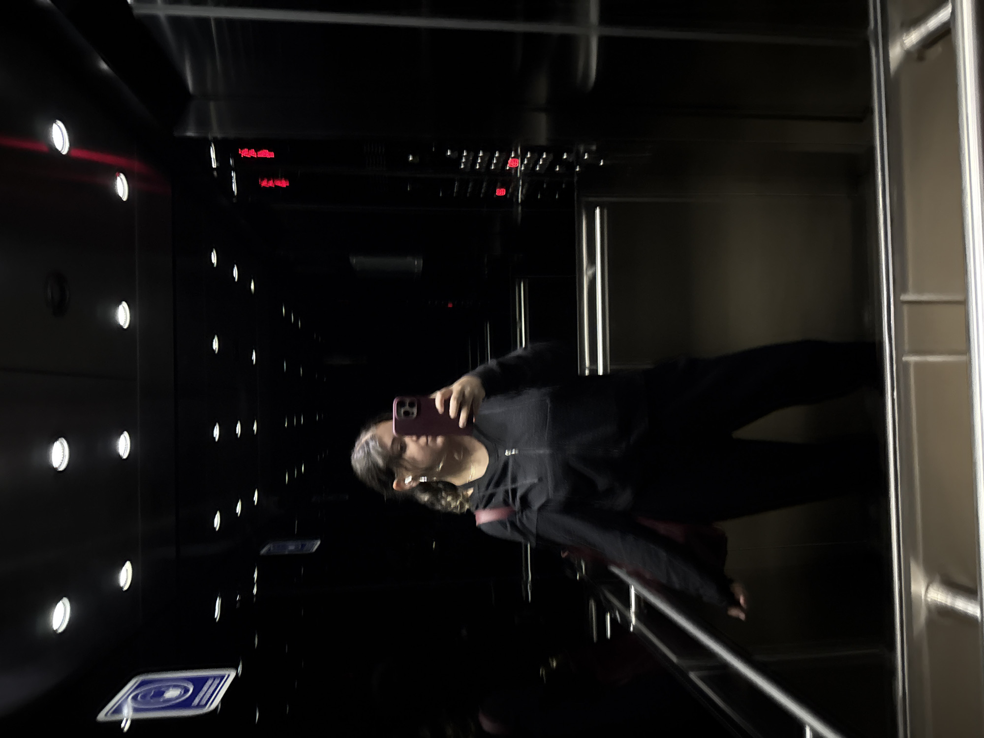 Here is a detailed description of the image:
**Scene:** An individual is taking a selfie inside what appears to be a modern elevator. The elevator features a dark interior with rows of illuminated push buttons and an electronic display showing numbers in red. The elevator doors have a metallic finish. There are handrails visible, suggesting a focus on accessibility and safety.
**People:** The image primarily features one individual holding a smartphone to capture a selfie. They are wearing dark clothing, possibly a sweatshirt and pants. The individual has dark hair and is positioned at an angle to the camera, likely to frame themselves within the reflection of the elevator's surfaces.
**Objects:** Key objects in the scene include the smartphone being used for the selfie, the elevator push buttons, the electronic display, the handrails, and signage on the floor.
**Actions:** The main action is the person taking a selfie in the elevator.
**Interactions:** The person is interacting with their phone, presumably to capture and possibly share the image. The presence of handrails suggests an interaction with the environment for support, though it is not directly observed.
**Environment:** The environment is a modern elevator interior. The style and materials suggest it could be located in a public or commercial building in Mexico City, Mexico. The details of the elevator interior, such as the push buttons, lighting, and signage, indicate a well-maintained and functional space.