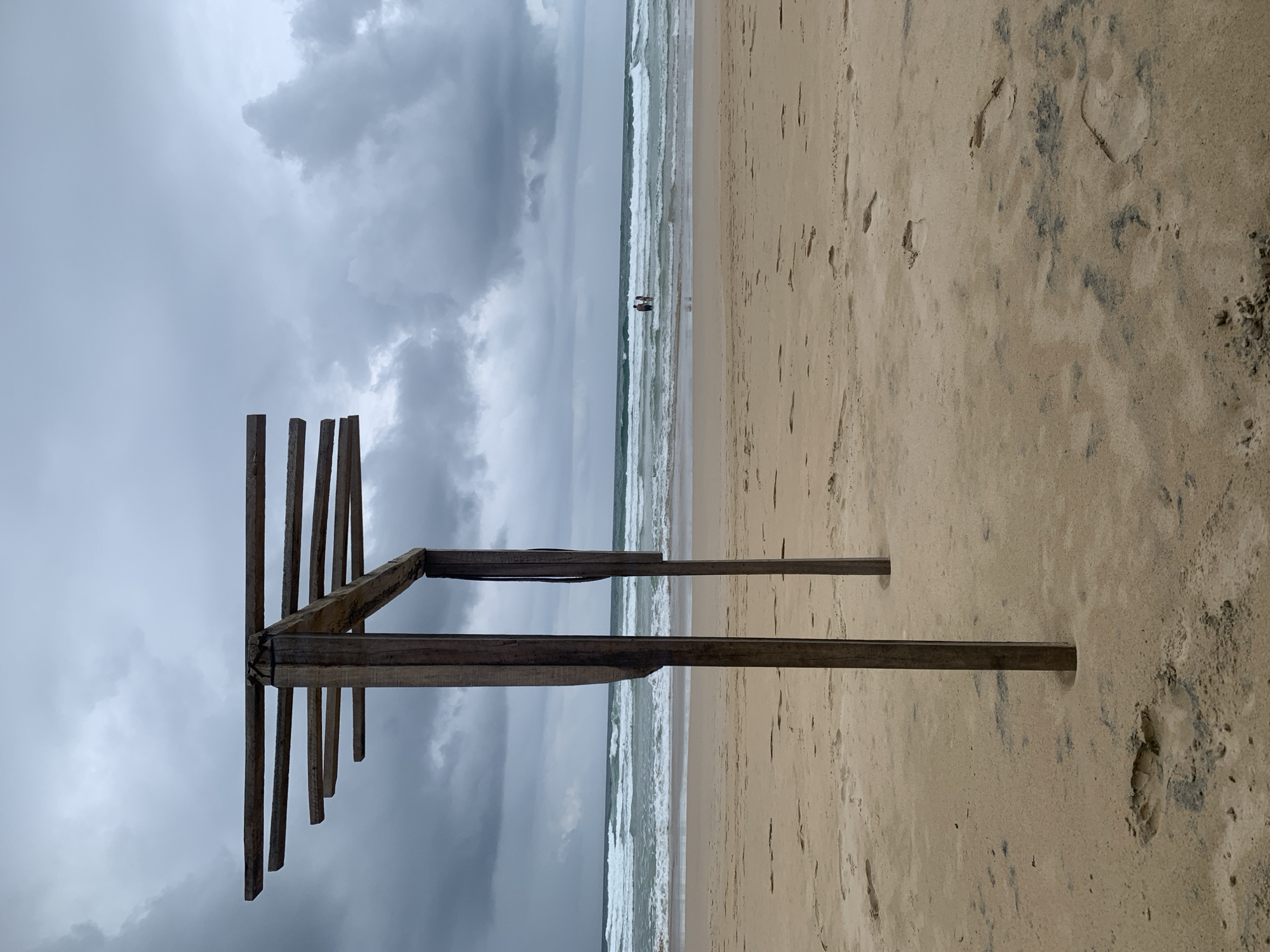 This is a coastal scene, likely a beach, near Kathaluwa, Sri Lanka. The photo depicts a sandy beach under an overcast sky with a view of the ocean. A weathered, wooden structure resembling a frame or dilapidated shelter is prominent in the foreground. Two figures are visible in the distance at the ocean's edge, possibly wading or swimming. The sand shows numerous footprints.