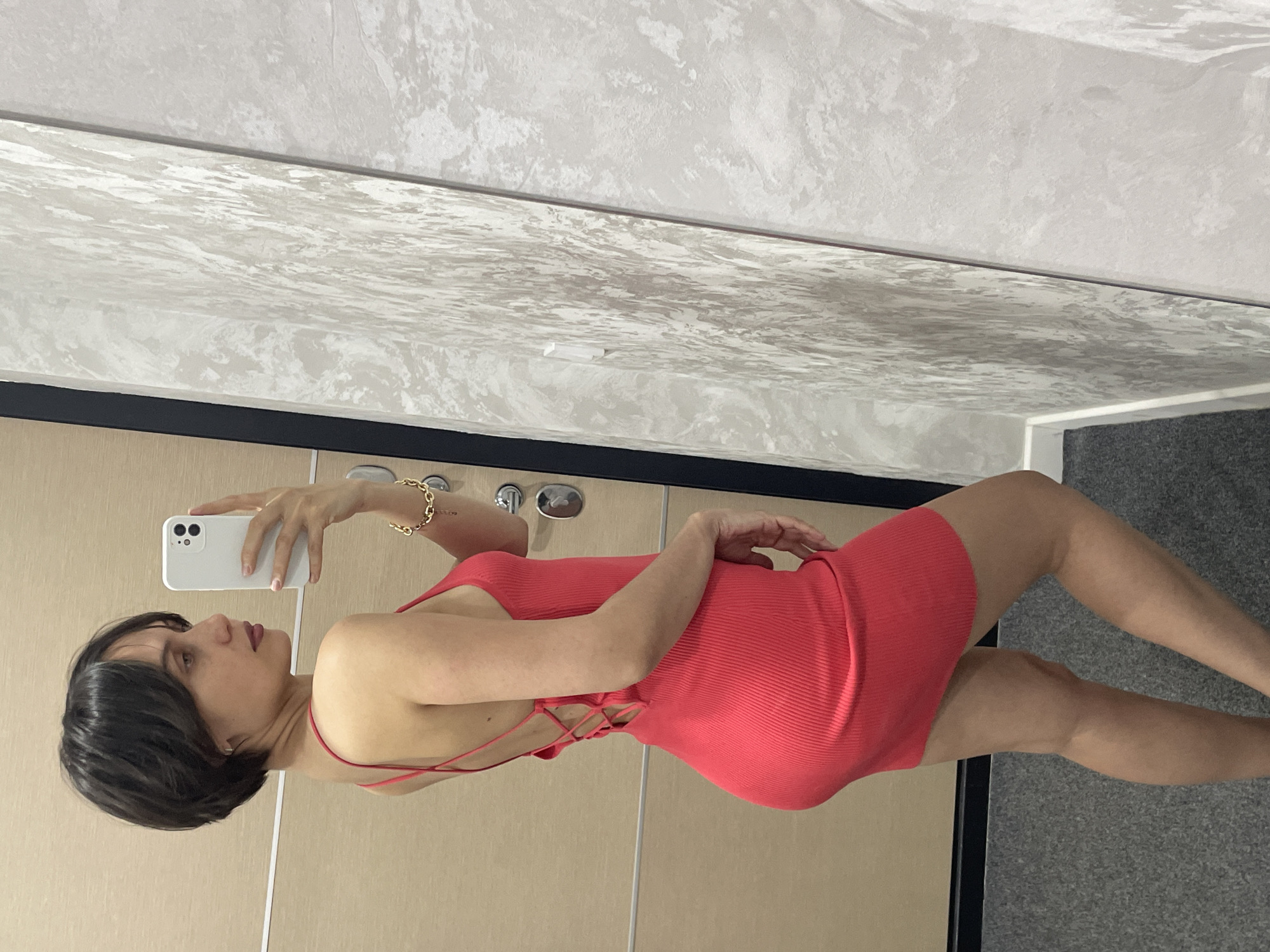 A woman with short, dark hair and fair skin is taking a selfie in what appears to be a dressing room or bathroom. She is wearing a tight, red dress with thin straps that crisscross at the back, and a gold bracelet on her left wrist. She is holding a white smartphone in her left hand, aimed at her reflection in a large mirror. The mirror reflects a textured, possibly marble or faux-marble wall. The background includes what looks like wooden paneling or doors. There are two round, metallic knobs or fixtures near the top edge of the paneling behind her. The location is indeterminable and cannot be said to be in Izhevsk, Russia based on the image provided.