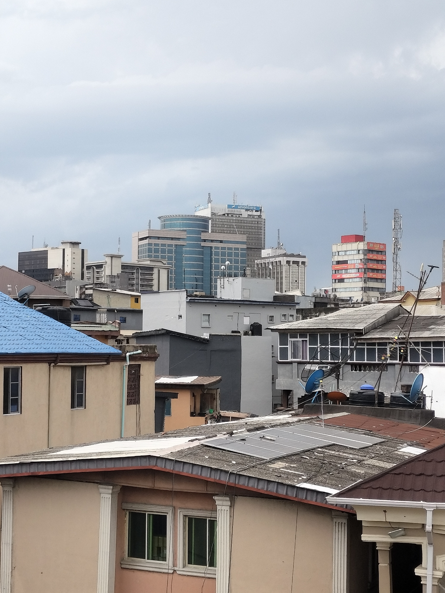 The image depicts a cityscape in Lagos, Nigeria, under an overcast sky. The foreground is dominated by residential buildings of varying heights and architectural styles, some with blue or brown roofs. Some buildings appear to have solar panels and satellite dishes. In the background, taller commercial buildings are visible, including one with "Unionbank" on top. The overall impression is of a densely populated urban area with a mix of residential and commercial structures.