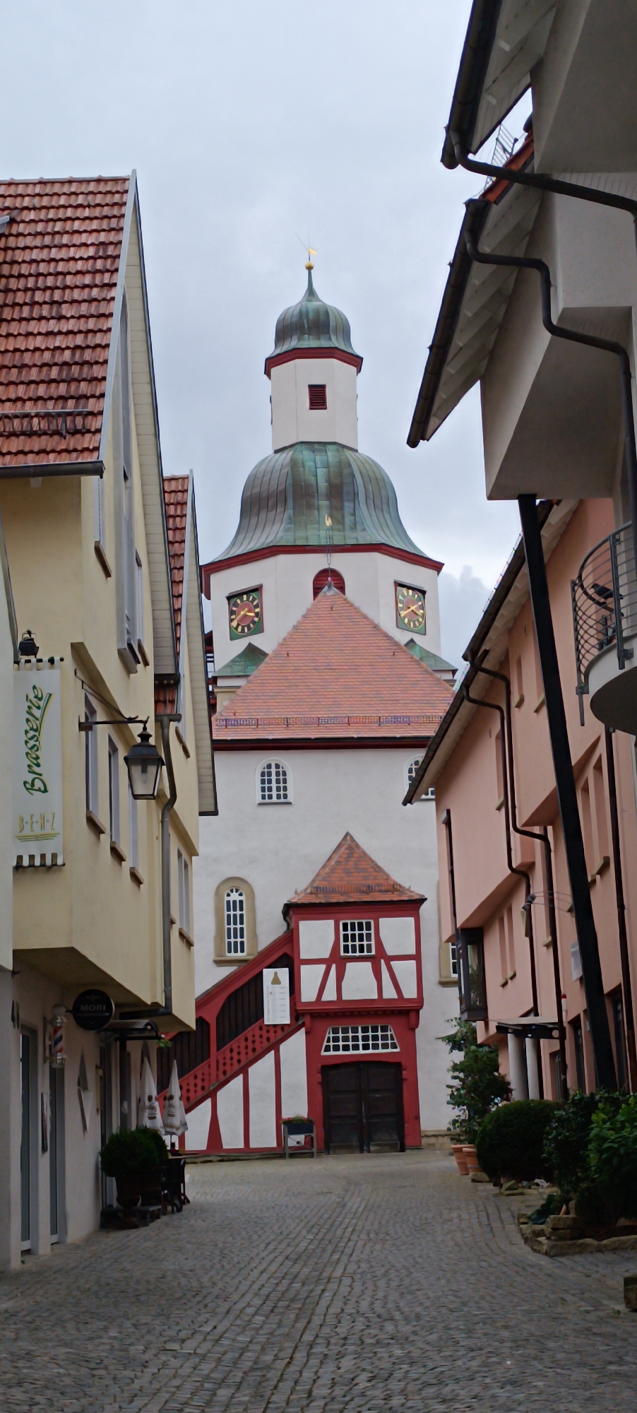 The image shows a view looking down a cobblestone street in Winnenden, Germany, towards a white church with a green dome. The church has clocks on its tower and a red-roofed building with white trim attached to its base. On the left side of the street is a pale yellow building with a sign that reads "Brasserie" and another sign for "MORI". On the right side of the street are several pink buildings with balconies and some potted plants. The sky is overcast. There are no people visible in the image.