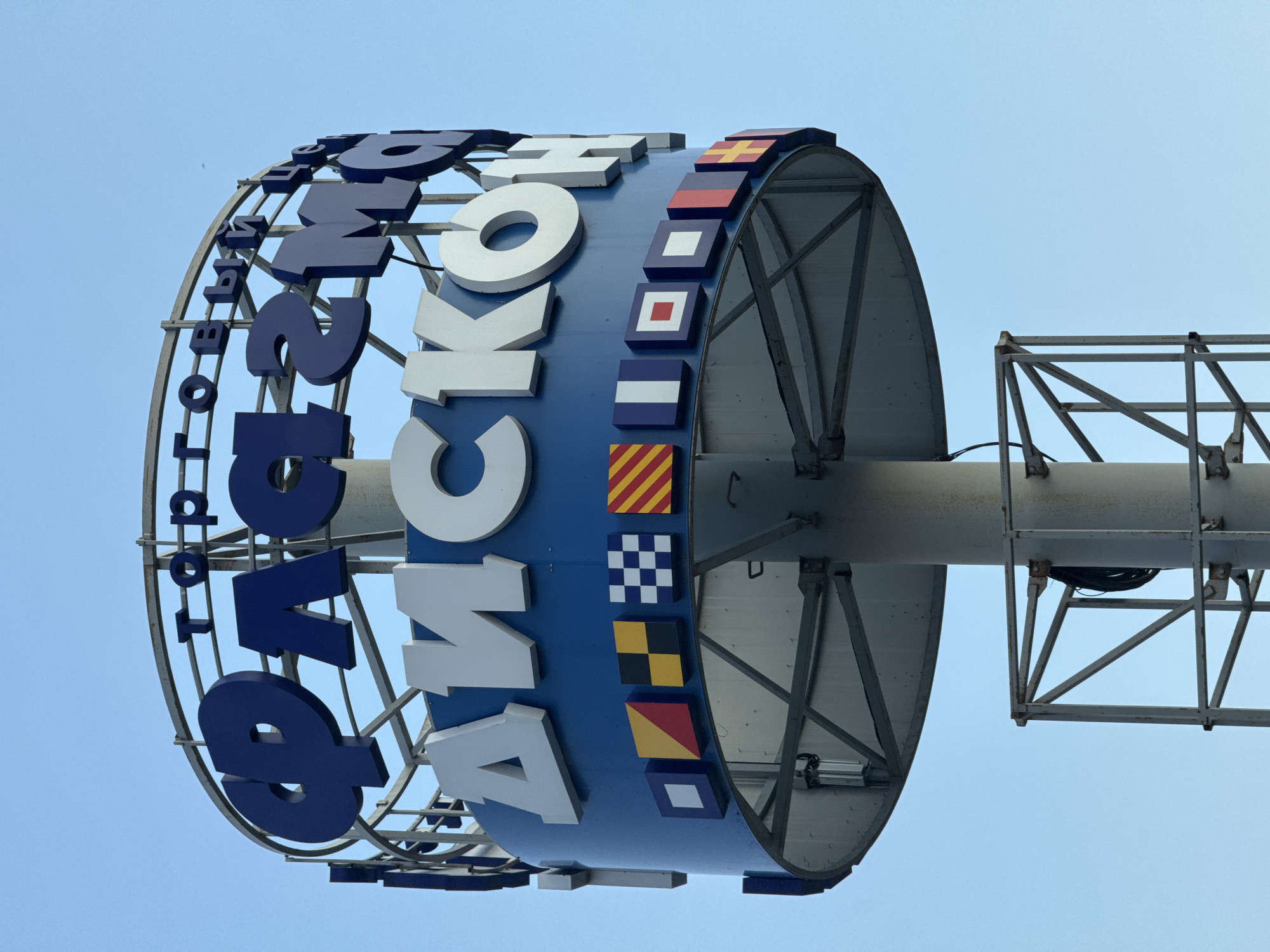 A large, cylindrical sign displaying Cyrillic letters and nautical flags is mounted on a metal pole against a clear, pale blue sky, suggesting daytime. The sign's prominent feature is the large, three-dimensional letters, predominantly dark blue and white, that form words around its circumference. The letters are angled, with some appearing to curve inward or outward, creating a dynamic visual effect.
The sign is decorated with a series of small, rectangular panels depicting various nautical flags. These flags have distinct patterns and colors, including red and yellow stripes, a checkered blue and white pattern, a yellow and black diamond, a red and yellow quartered flag, and a solid white diamond on a blue background. These flags are arranged vertically along a blue section of the sign.
The sign is supported by a complex metal structure, including a curved framework that forms the outer shell of the cylinder and a robust metal pole extending upwards and to the right. A secondary metal structure, resembling a scaffold or platform, is attached to the pole to the right of the sign. Some visible wires and electrical components are also present within the supporting structures.
The perspective of the photograph is from below, looking up at the sign. The sky is uniformly clear, indicating fair weather. The scene appears to be outdoors, likely in an area where a prominent landmark or business is located, given the size and design of the sign. The text on the sign is in Russian. The word "ТОП" is visible on the left, and "ЛОГ" appears to the right of it, with "НОВ" further to the right. The white letters in the center appear to spell "Я КОН".