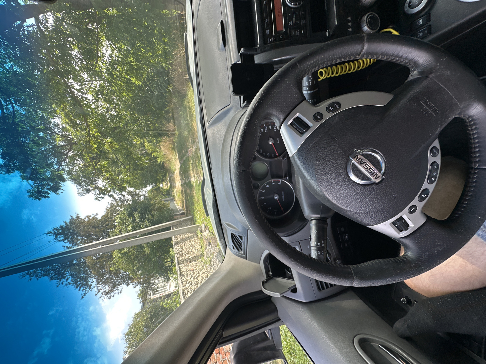 The image is a first-person view from inside a Nissan vehicle, looking forward through the windshield and at the dashboard. The vehicle appears to be on a dirt or gravel road lined by trees and a stone wall. A power pole with electrical wires is visible in the distance, leaning slightly to the left. The sky is blue with some scattered clouds.
The dashboard features a steering wheel with the Nissan logo, various buttons, and a speedometer. A phone mount is attached to the center of the dashboard. To the right, a car radio is visible. The interior of the car shows grey plastic and leather upholstery. The driver's left leg, clad in a black sock, is visible on the left side of the frame.
The location is identified as Makiv, Ukraine, and the scene suggests the vehicle is driving in a rural or semi-rural area outside of the main town. The lighting indicates it is daytime, likely mid-morning or afternoon, given the bright sunlight and clear sky.