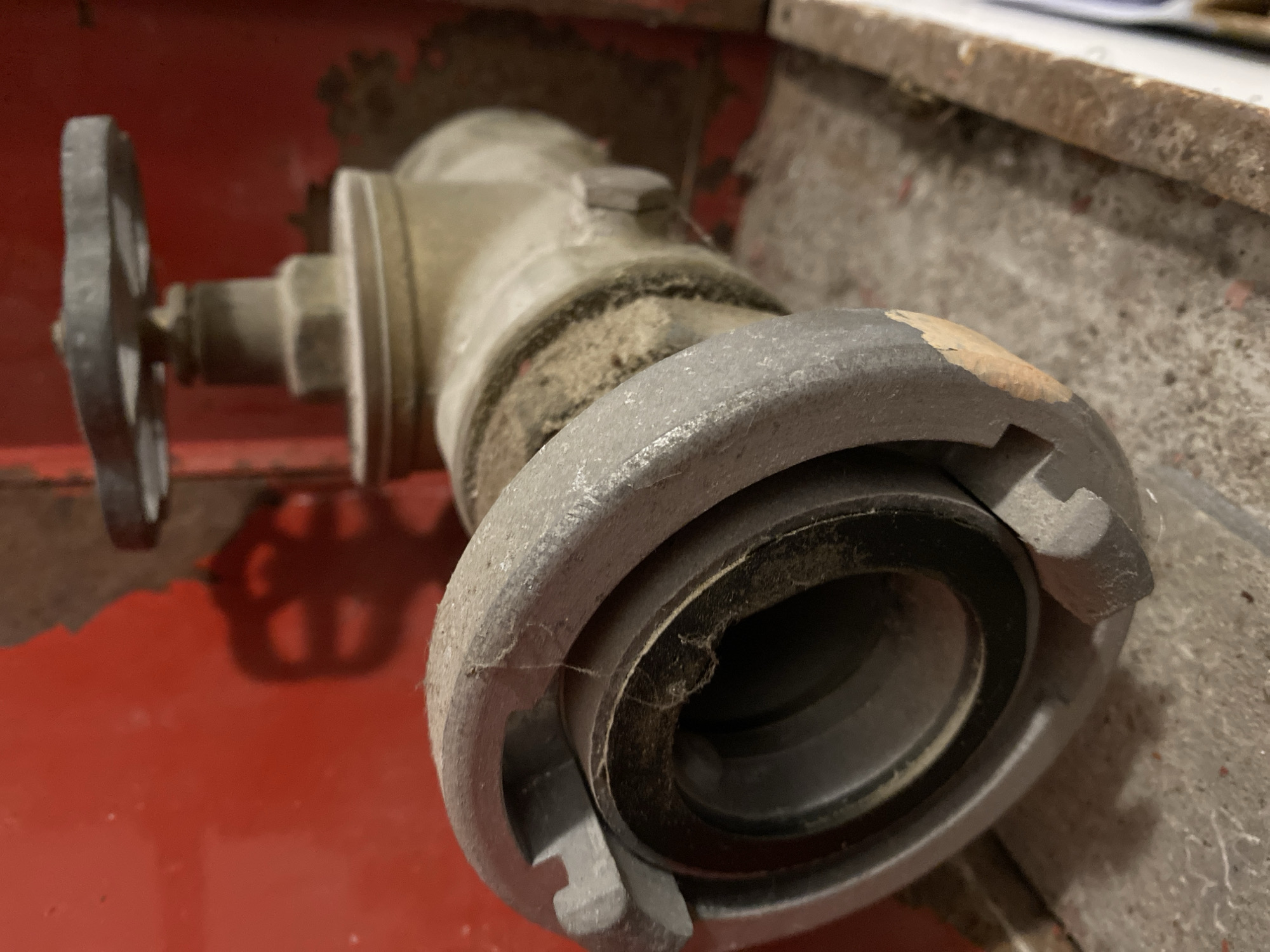 The image depicts a dusty, metallic valve or hydrant, possibly a fire hydrant connection, mounted on a red surface. It appears to be indoors or in a confined space. The valve is cylindrical, with a round handle on the left side and a connector on the right, showing visible signs of wear and dirt. There are traces of spiderwebs, indicating it has been undisturbed for some time. There is no evidence to suggest a specific location in Budva, Montenegro. The image primarily showcases the static object and its condition. There are no people or actions depicted.