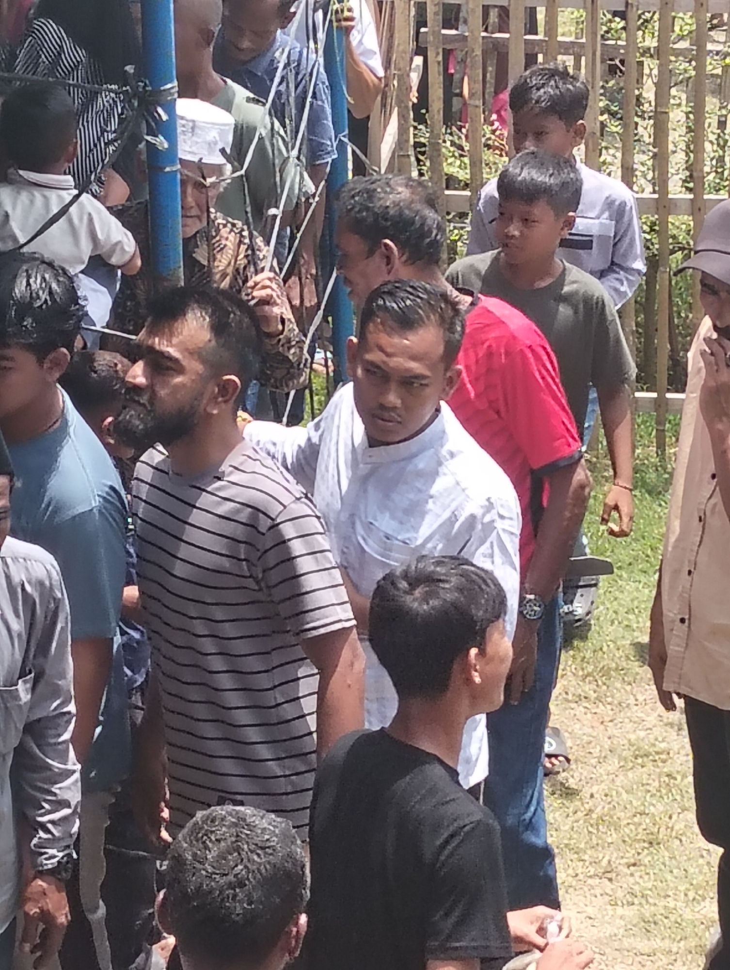 The image depicts a group of people gathered outdoors, with a bamboo fence in the background and dry grass on the ground. The scene appears to be in Grong-Grong, Indonesia. In the foreground, several individuals are visible. A man wearing a striped t-shirt with black and grey lines is looking to his right. To his left, a man in a light blue t-shirt is also present. Further to the left, the back of a person in a grey shirt is visible. Behind the man in the striped shirt, a person in a white shirt is looking forward. A man in a bright red shirt is to the right of the person in the white shirt. In the background, other individuals are standing, including a child in a light blue shirt and another child with dark hair. An older man wearing a white cap and a patterned brown and white garment is partially obscured by a blue pole. The lighting suggests it is daytime.