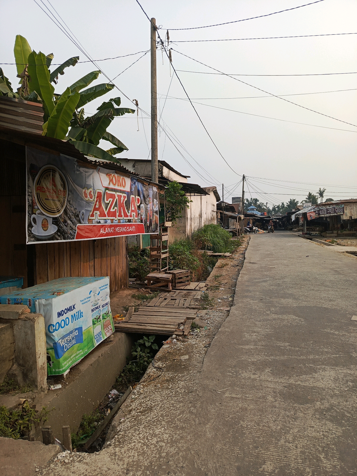 The image depicts a street scene in Megang Sakti I, Indonesia. On the left side of the street, there is a storefront with a banner that reads "TOKO AZKA" and "ALAMAT: MEGANG SAKTI". In front of the storefront, there are several boxes of "INDOMILK GOOD Milk" stacked. A drainage ditch runs alongside the sidewalk, with some vegetation and debris visible. Wooden planks are laid across the ditch, forming a makeshift bridge. Further down the street, there are more buildings and stalls, suggesting a local market or a commercial area. A single utility pole with numerous overhead wires is prominent in the foreground. The street is paved with concrete and appears to be a well-trafficked area. The sky is overcast, and the overall lighting suggests it is daytime. There are no people clearly visible in the immediate foreground, but a person on a motorcycle is seen in the distance.