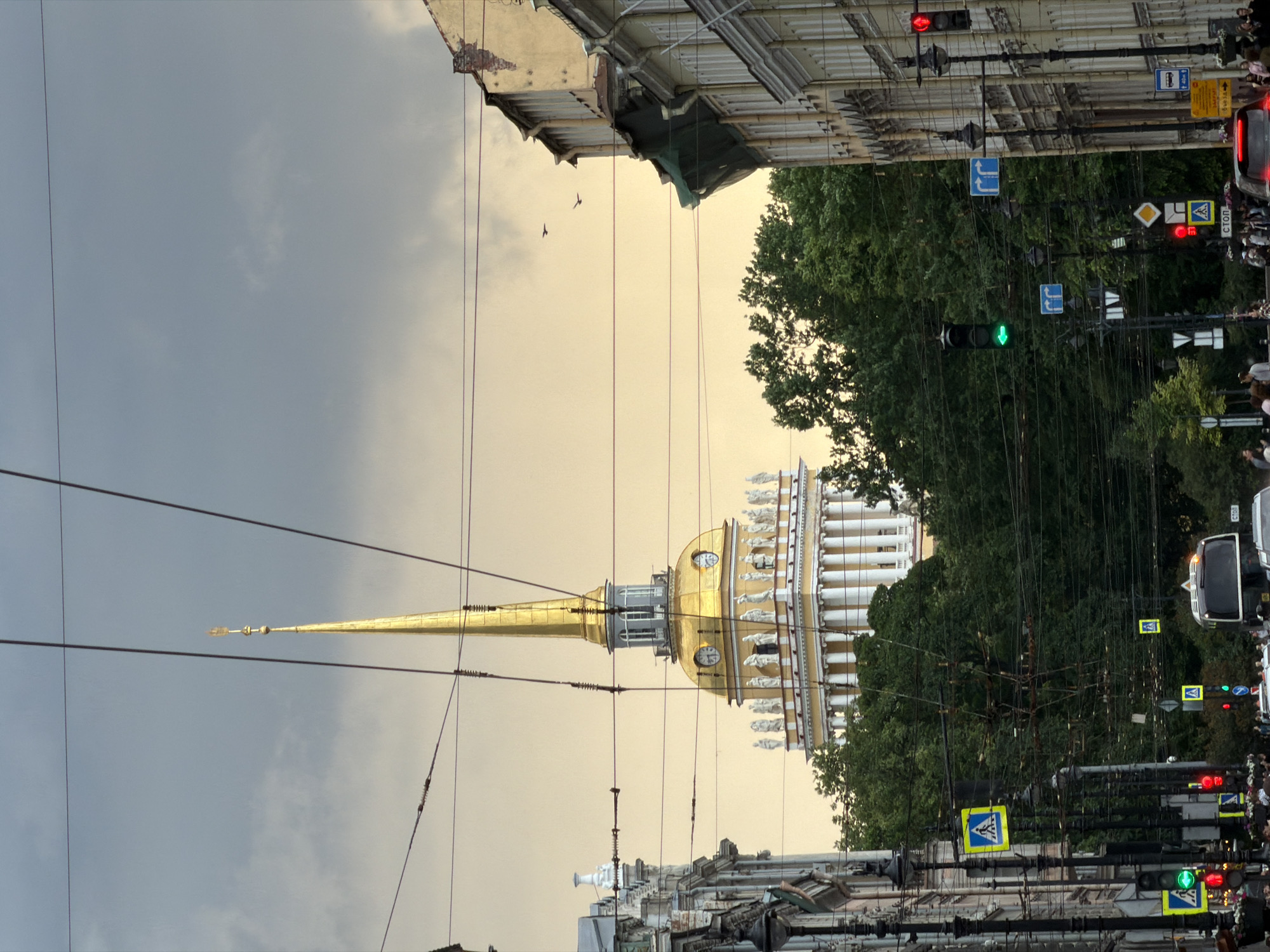 This image captures a view of the Admiralty building's spire in Saint Petersburg, Russia, framed by overcast skies and overhead wires. The iconic golden spire, topped with a weather vane, dominates the upper half of the frame. Below the spire, the building's white facade is adorned with statues and columns. Lush green trees partially obscure the lower portions of the building and extend to the right and left.
In the foreground, a street scene unfolds. Buildings line the right side of the street, their facades showing signs of age. Several streetlights and traffic lights are visible, with a green traffic light illuminated. Pedestrian crossing signs with blue backgrounds and white symbols are also present. A bus and other vehicles are on the street, along with a crowd of people walking on the sidewalk. Two birds are flying in the sky on the right side of the image. The overall atmosphere suggests a somewhat muted, possibly late afternoon or early evening light due to the cloudy conditions.